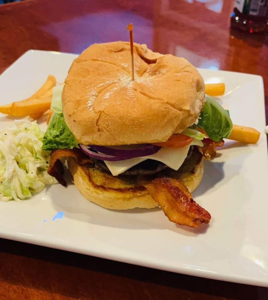 Hamburger with bacon, lettuce, tomato, and red onion on a white plate with fries and coleslaw.