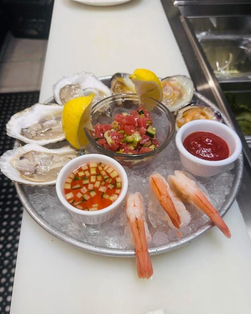 Seafood platter on ice: oysters, clams, shrimp, sauce, and lemon wedges.