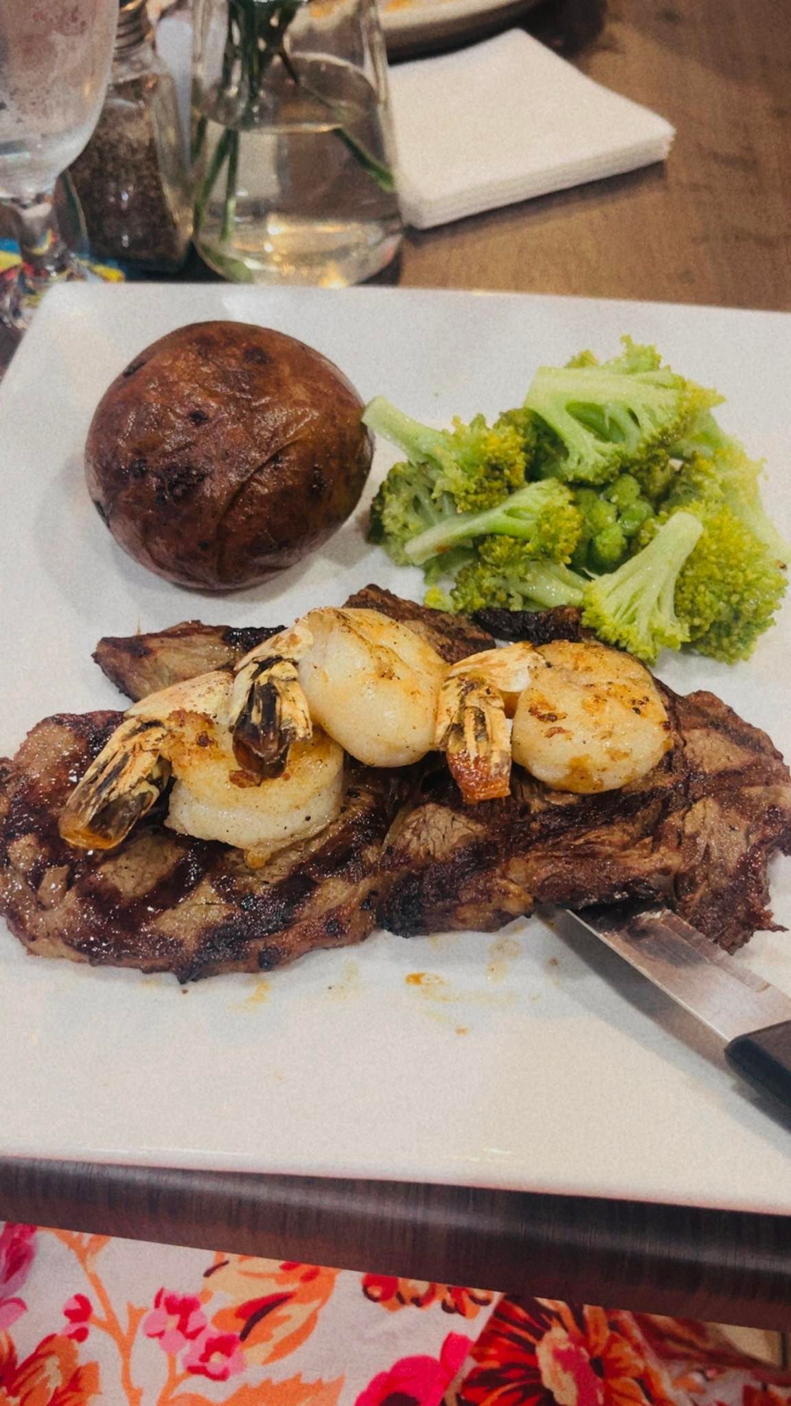 Steak topped with grilled shrimp and scallops, baked potato, broccoli on white plate.