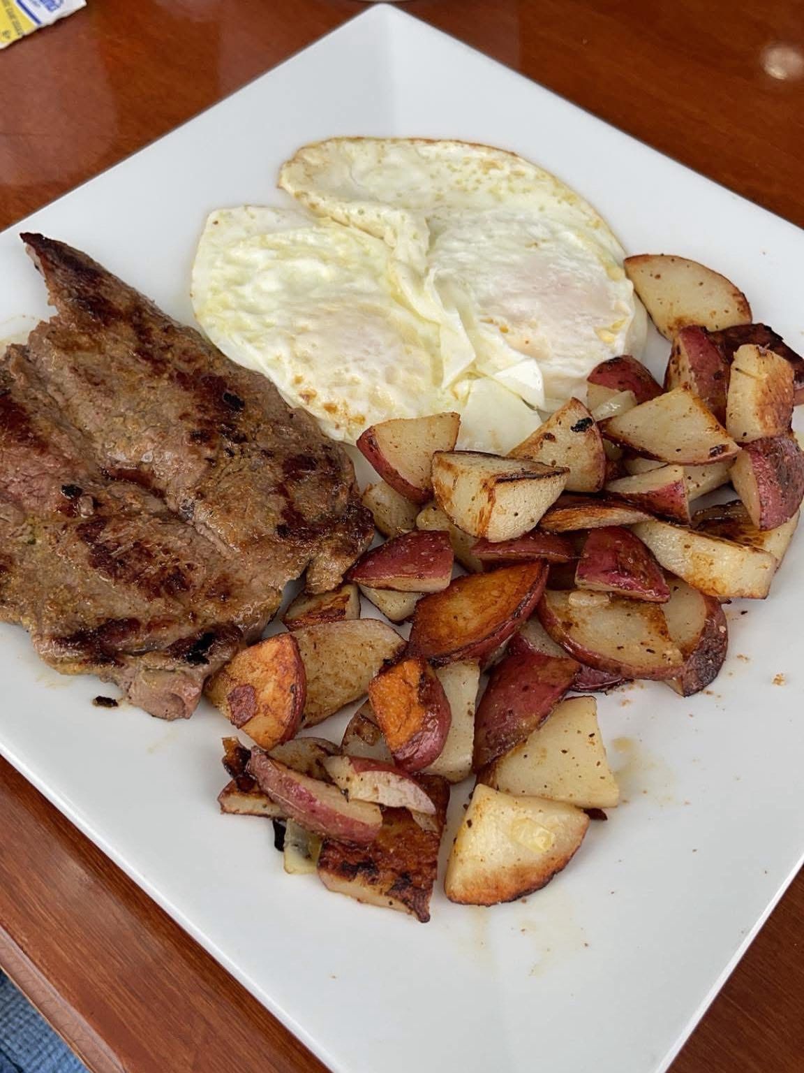 Steak, two fried eggs, and seasoned red potatoes on a white square plate.