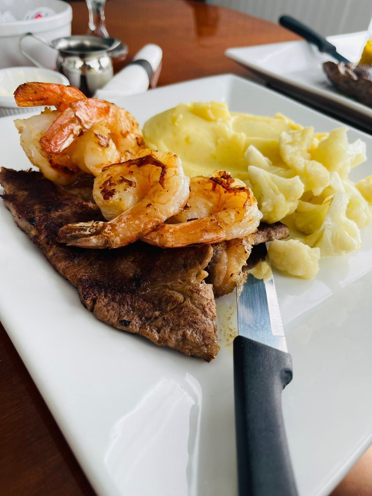 Steak topped with shrimp, served with mashed cauliflower and a side of cauliflower on a white plate.