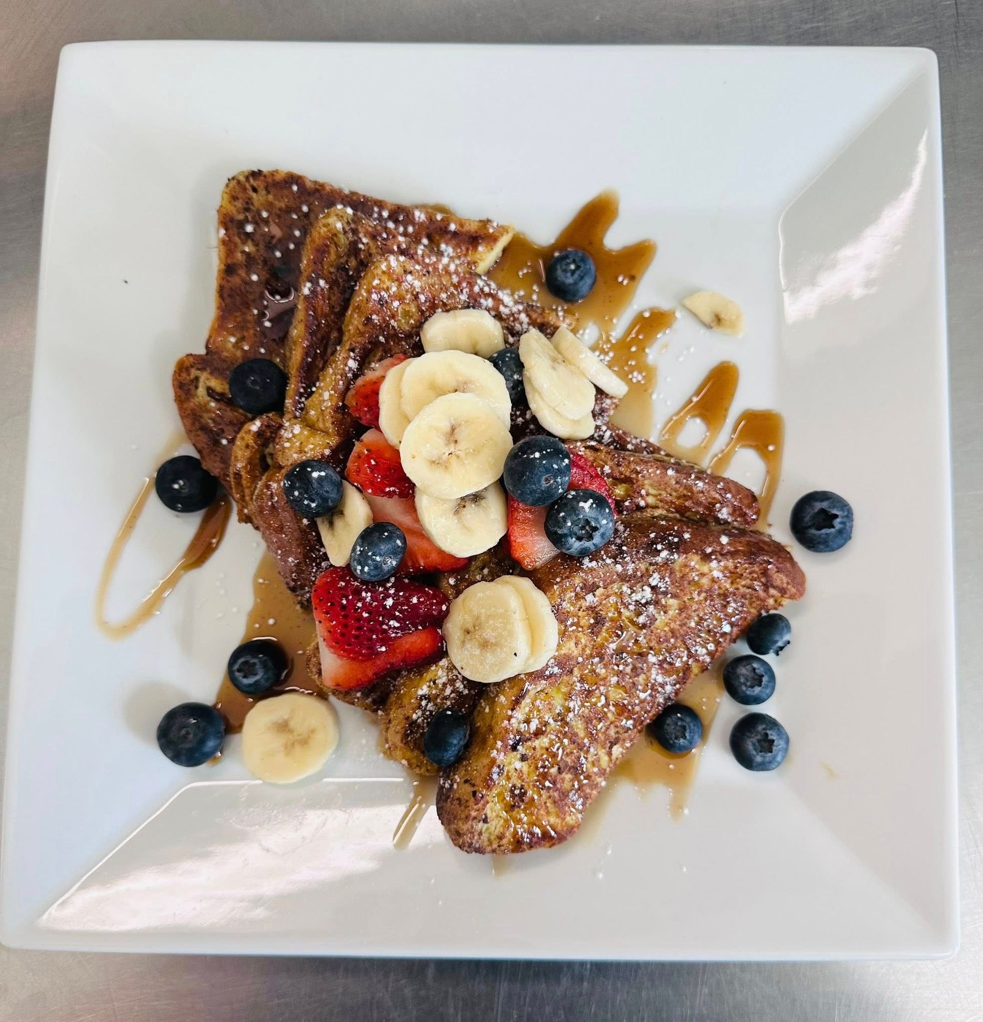 French toast with blueberries, strawberries, bananas, and syrup on a white plate.