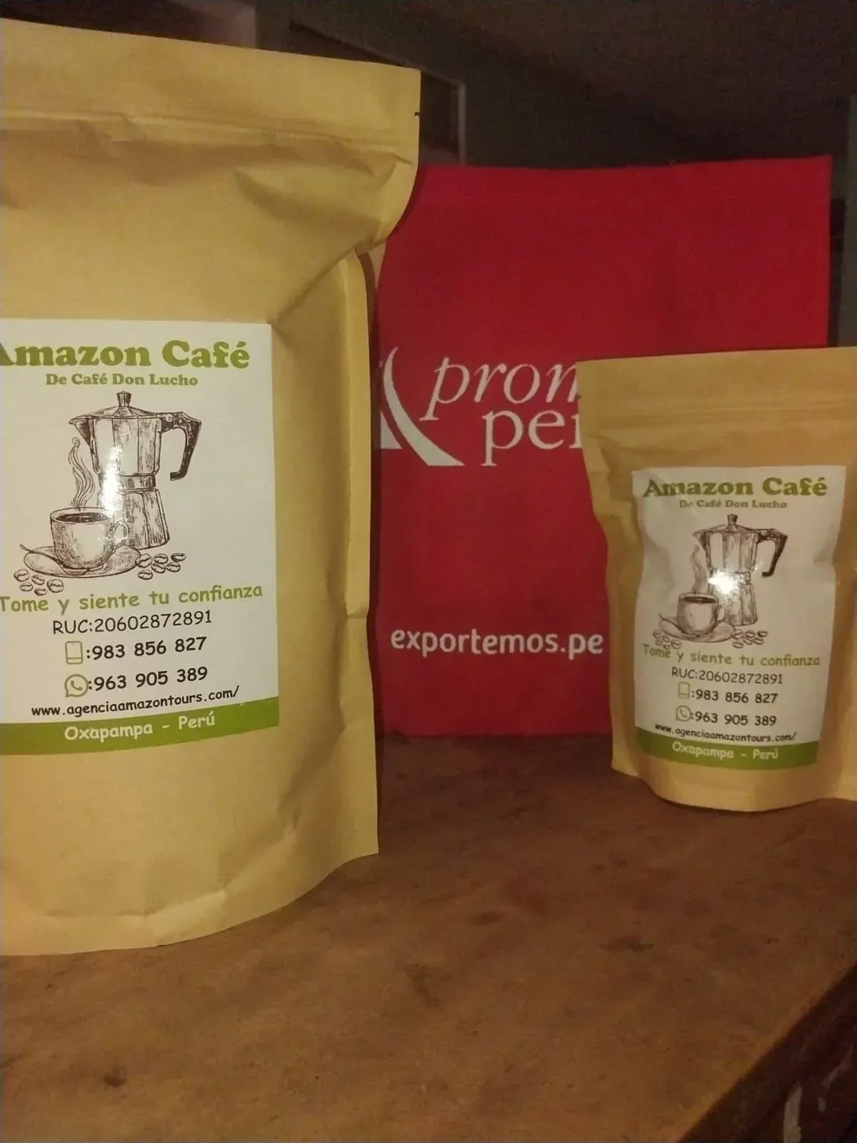 Bolsas de Amazon Café y un pequeño tarro sobre una mesa de madera, con una bolsa roja de Exportsmo.pe al fondo.
