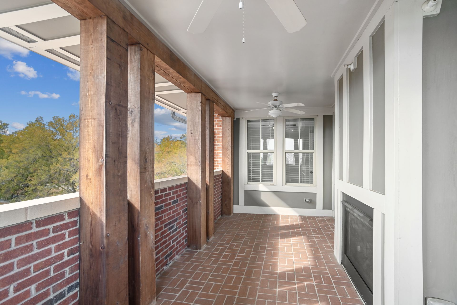 Covered outdoor patio with exposed wooden beams, brick flooring and wall, fireplace, and window.