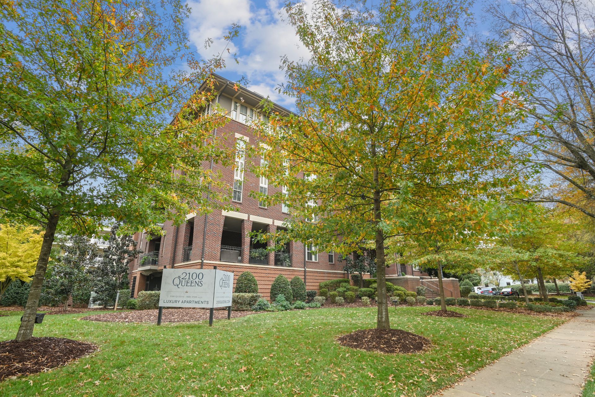Brick townhome behind green lawn and trees, sign in foreground, sidewalk to the right.