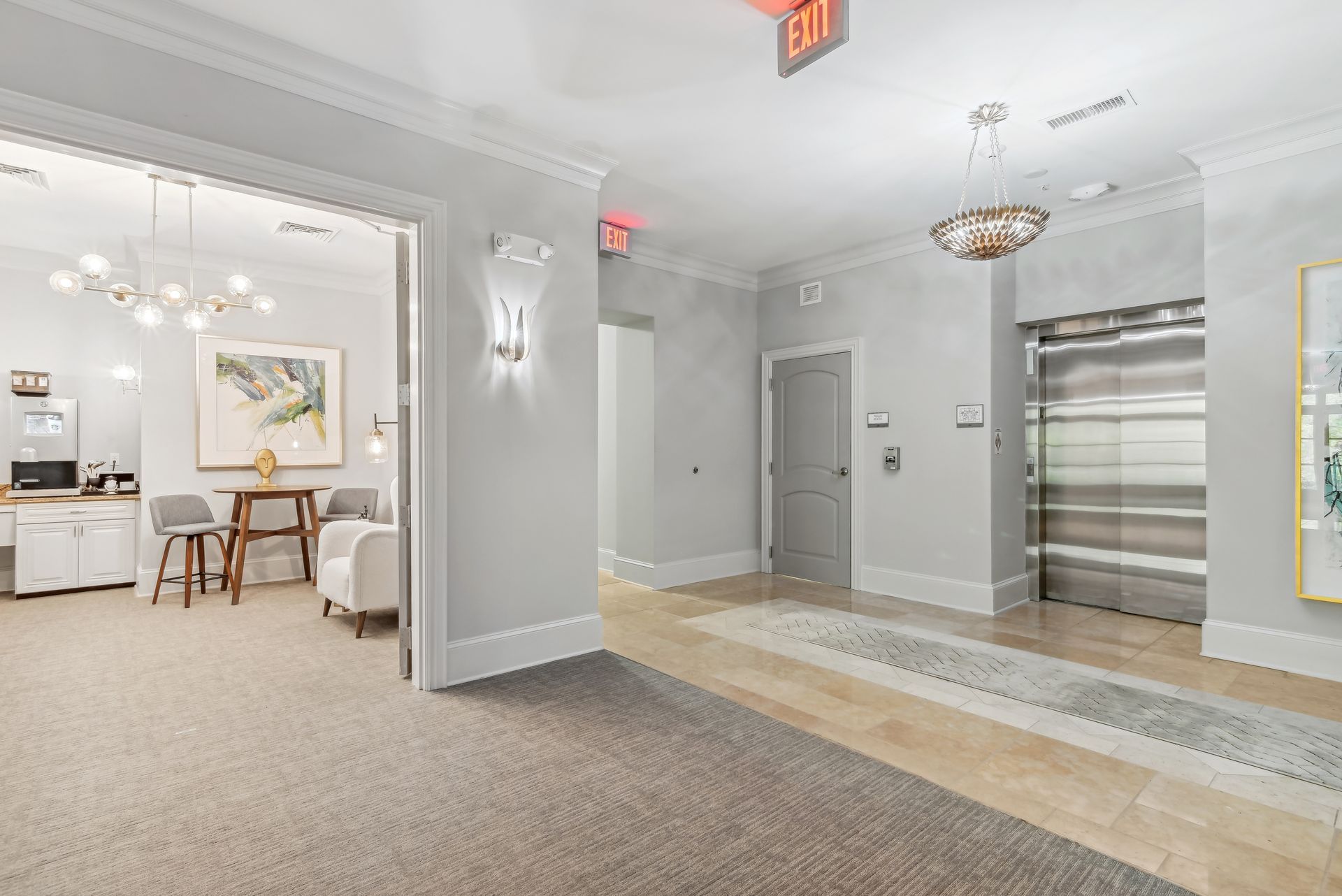 Lobby with open office area, elevator, and exit sign. Light gray walls, carpet, and neutral decor.