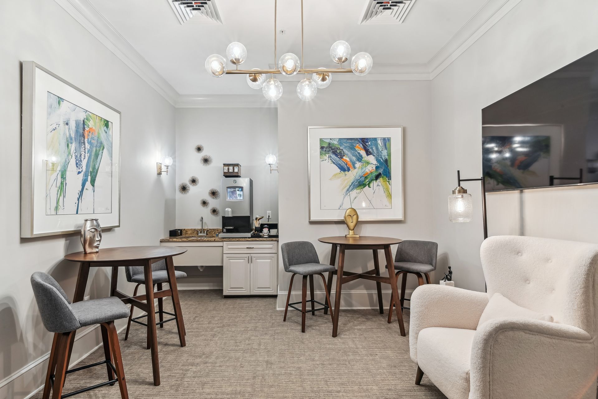 Cozy lounge area with bar tables, chairs, art, a sofa, and a light fixture, against neutral walls.