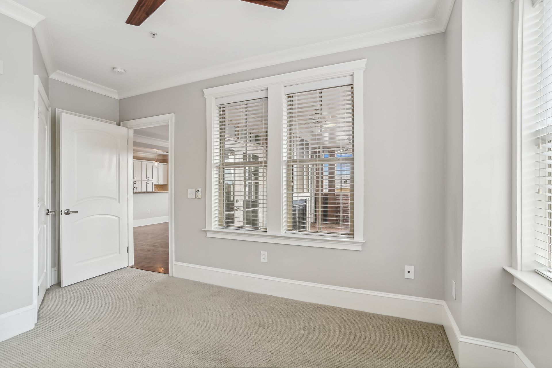 Empty room with a large window, white door, and beige carpet. Light gray walls and white trim.