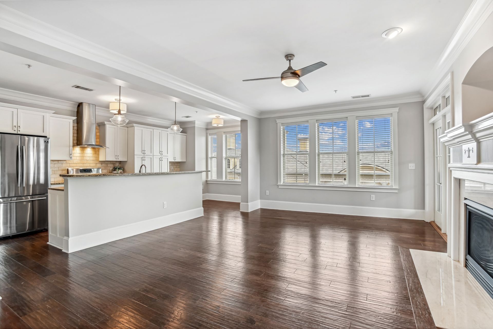 Open-concept living space with kitchen and fireplace. White walls, dark wood floors, large windows.