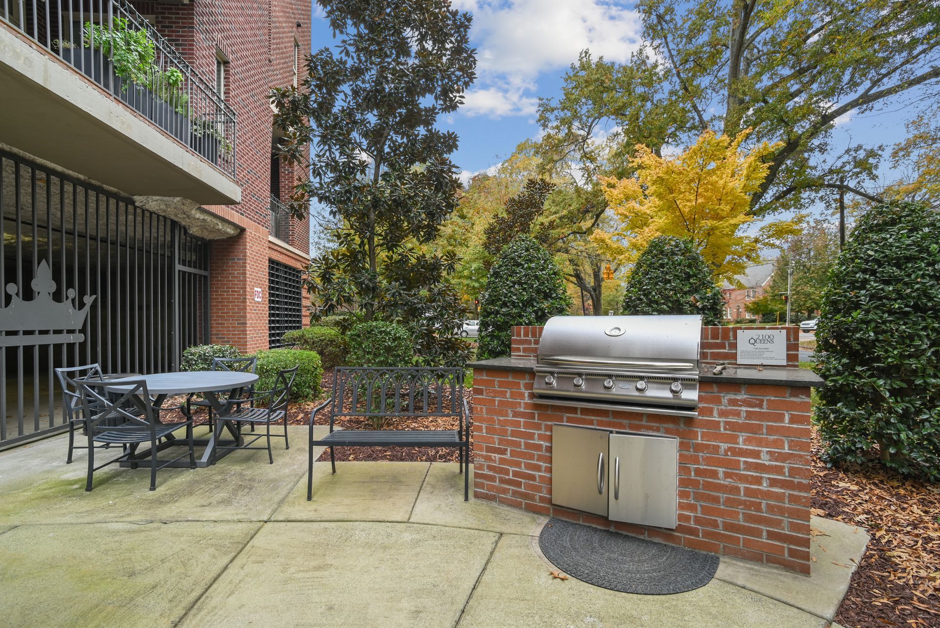 Brick patio with grill, table, bench, and greenery. Building in background.