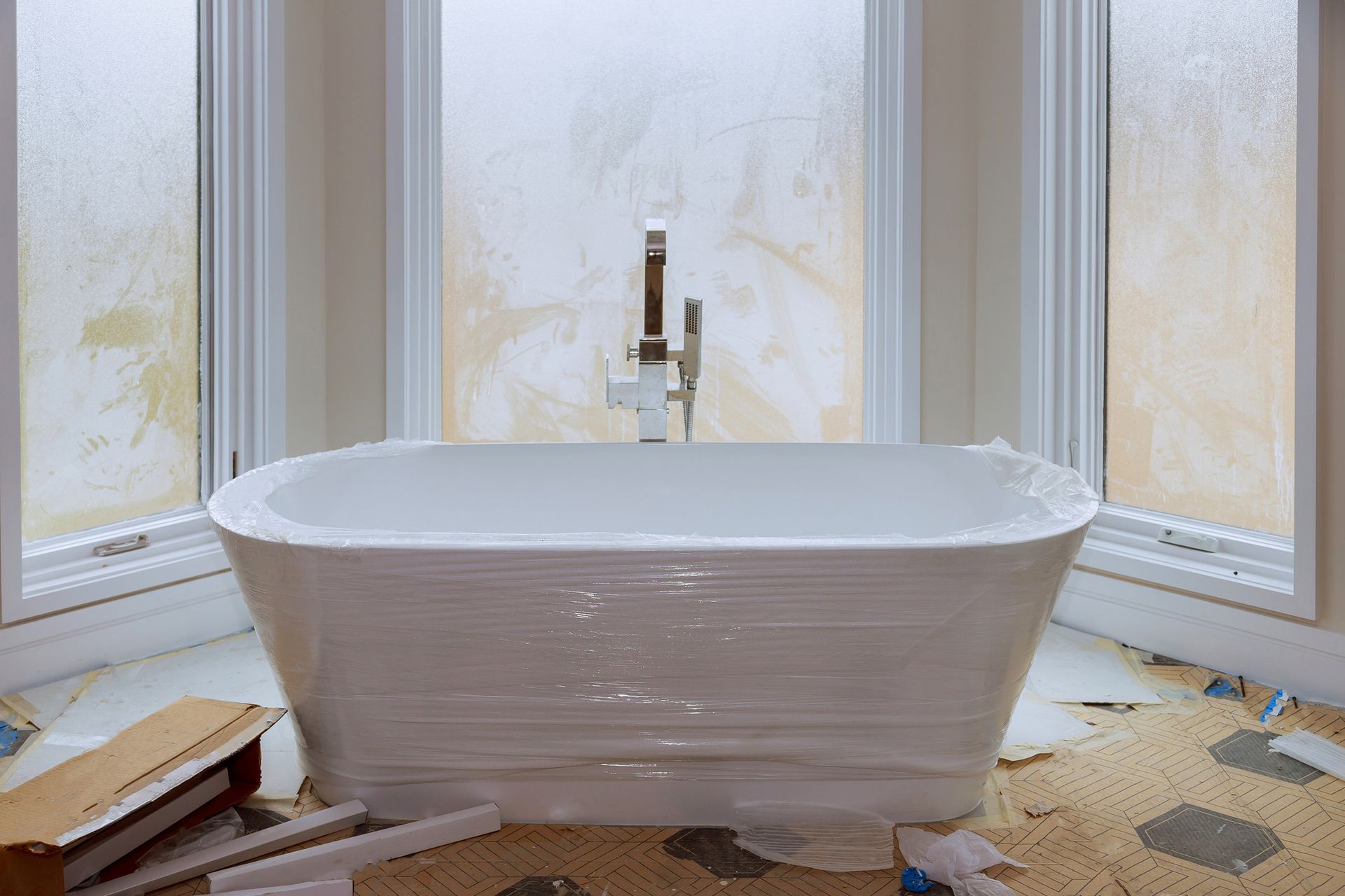 White bathtub wrapped in plastic in front of a large window. Construction debris on the floor.