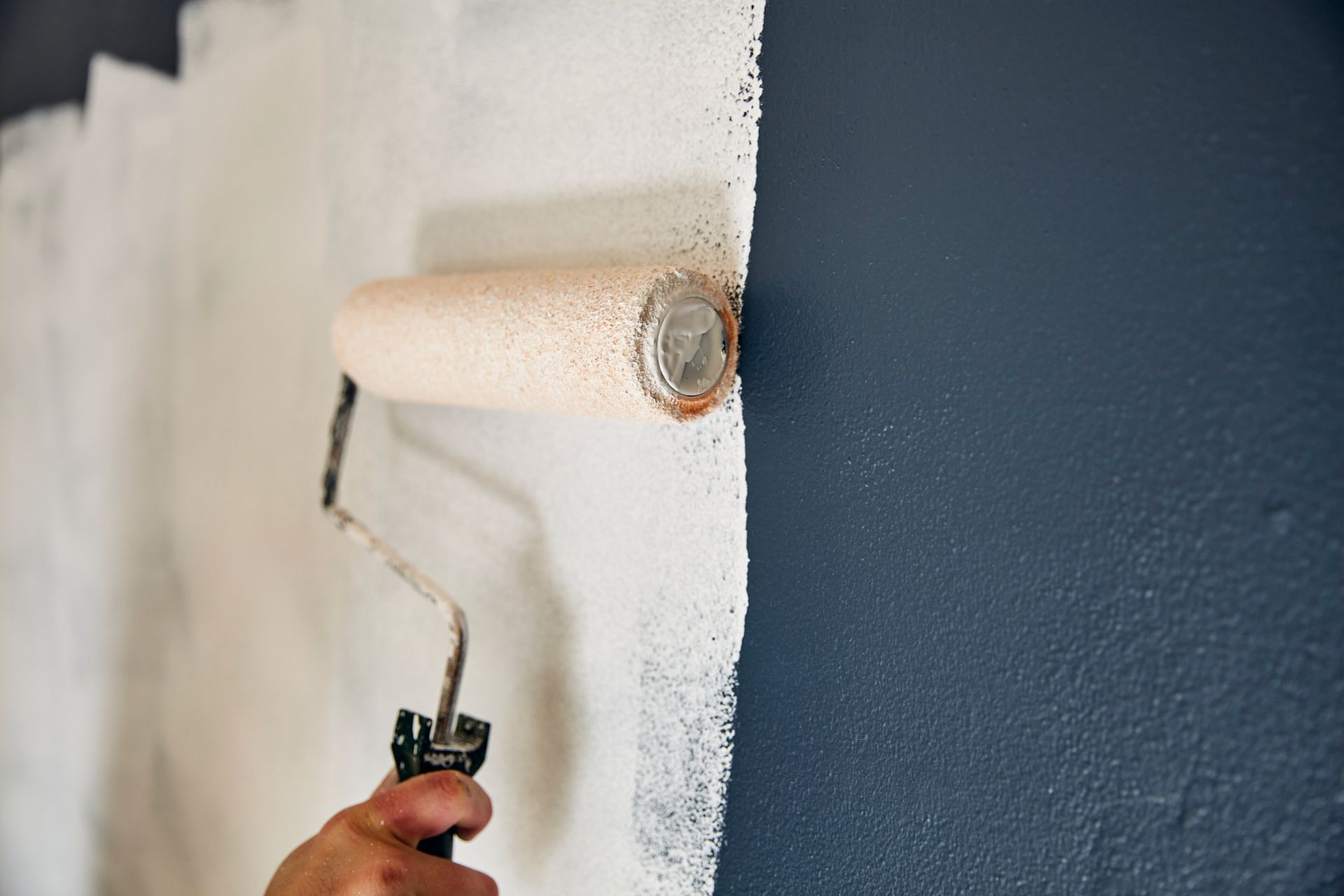Person using paint roller to apply white paint to a dark blue wall.