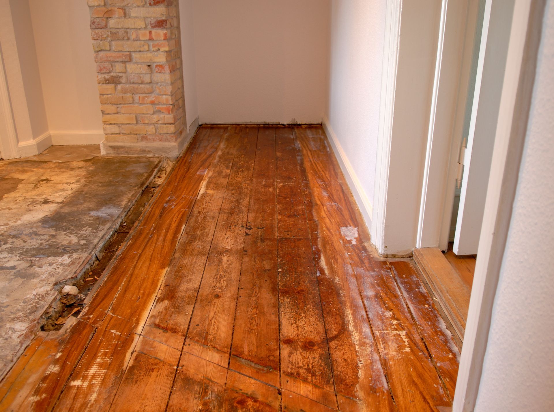 Wooden floor with sections of exposed concrete, next to brick pillar and white walls.