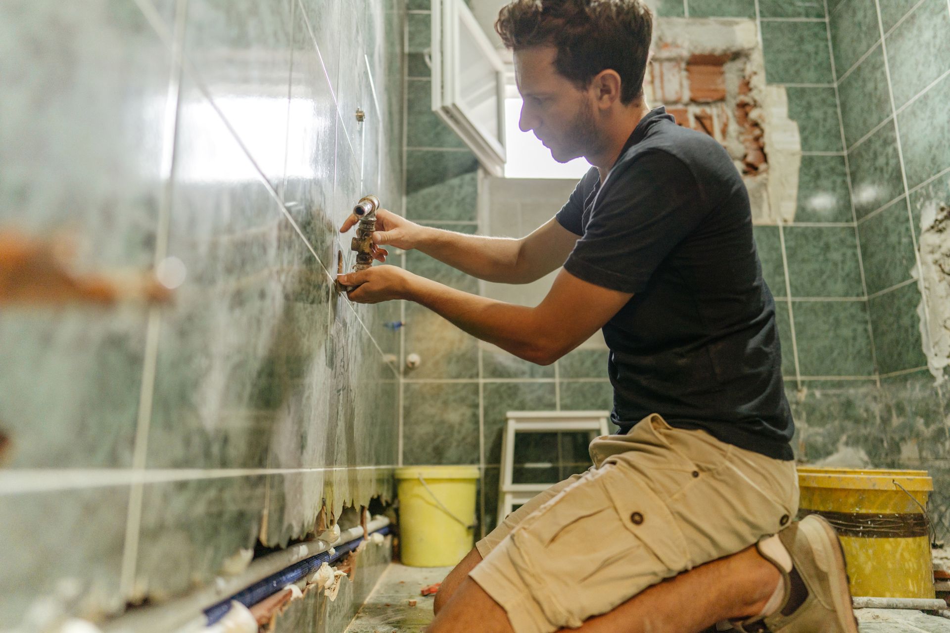 Man kneels in a bathroom, working on plumbing. Green tiles, exposed brick, and a yellow bucket are visible.