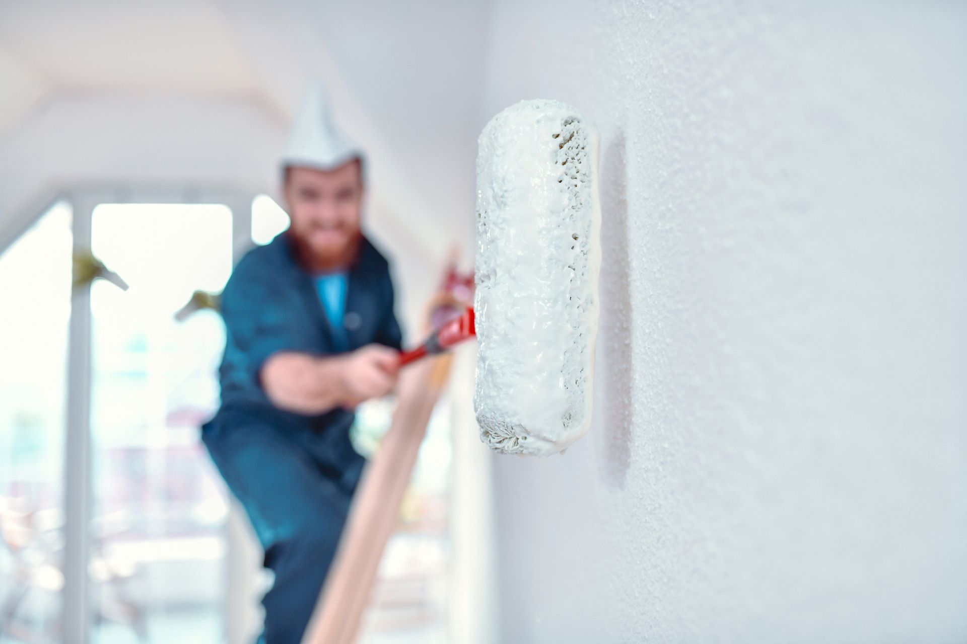 Painter on a ladder using a roller to paint a white wall indoors.