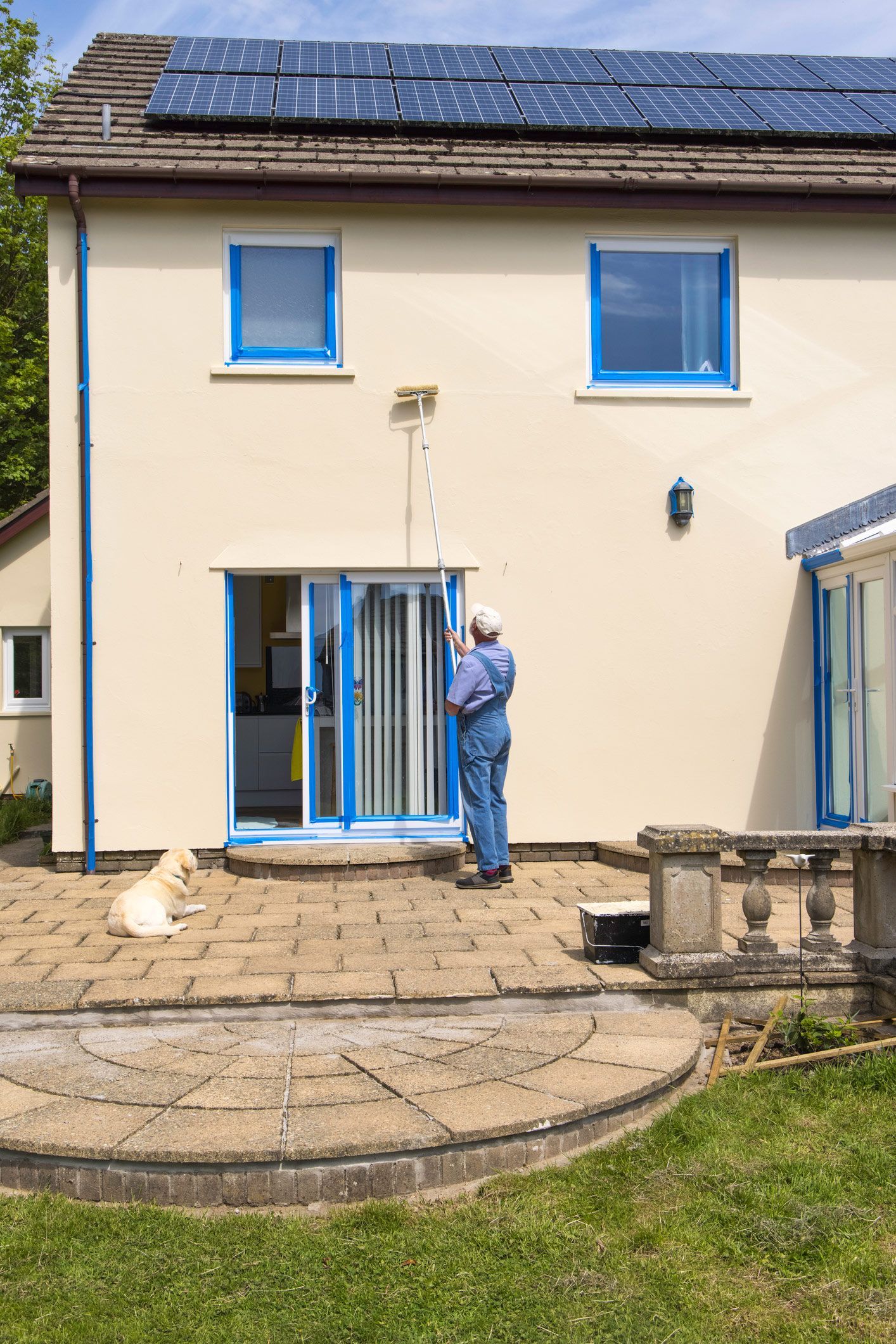 A person in overalls painting a blue window frame on a light yellow house with solar panels. A dog sits on the patio.