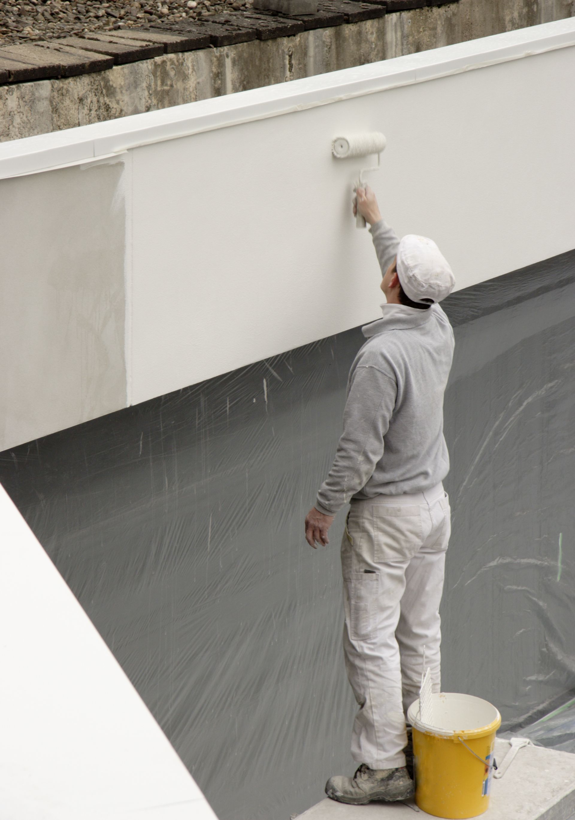 Person in white overalls painting a white wall with a roller, standing on a black surface.