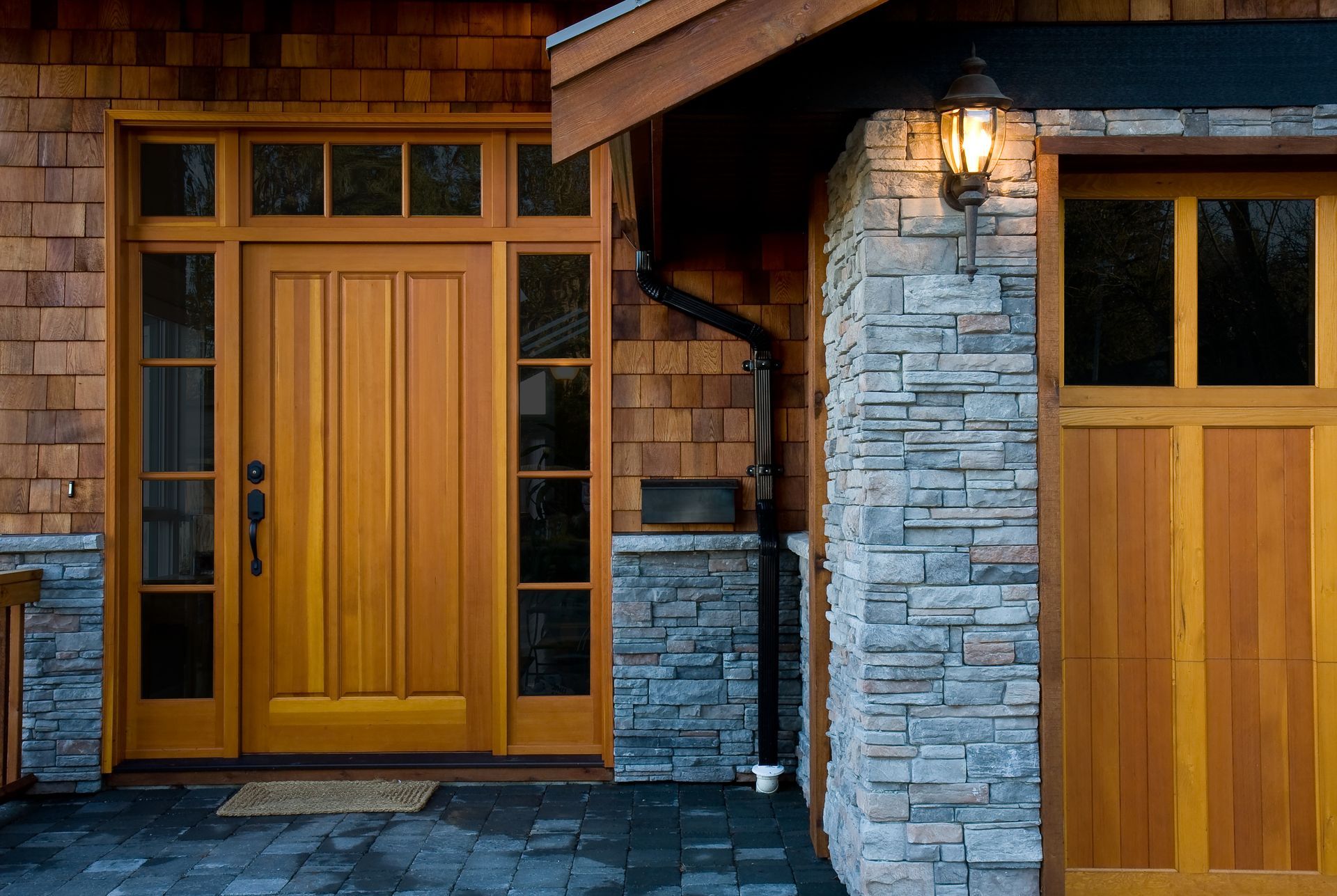 Wooden front door and garage door with stone accents, illuminated by a sconce.