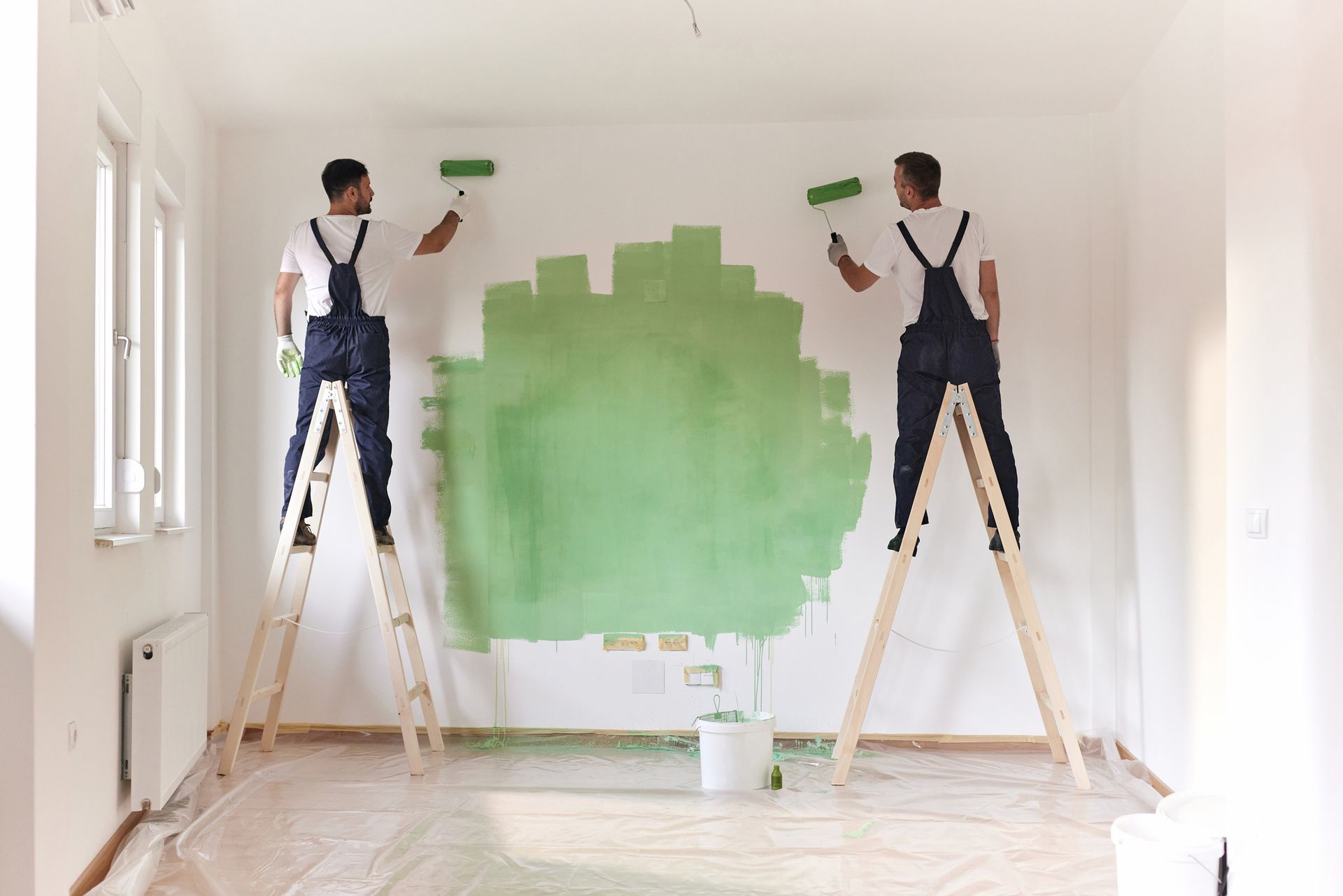 Two men painting a wall green while standing on wooden ladders in a room.