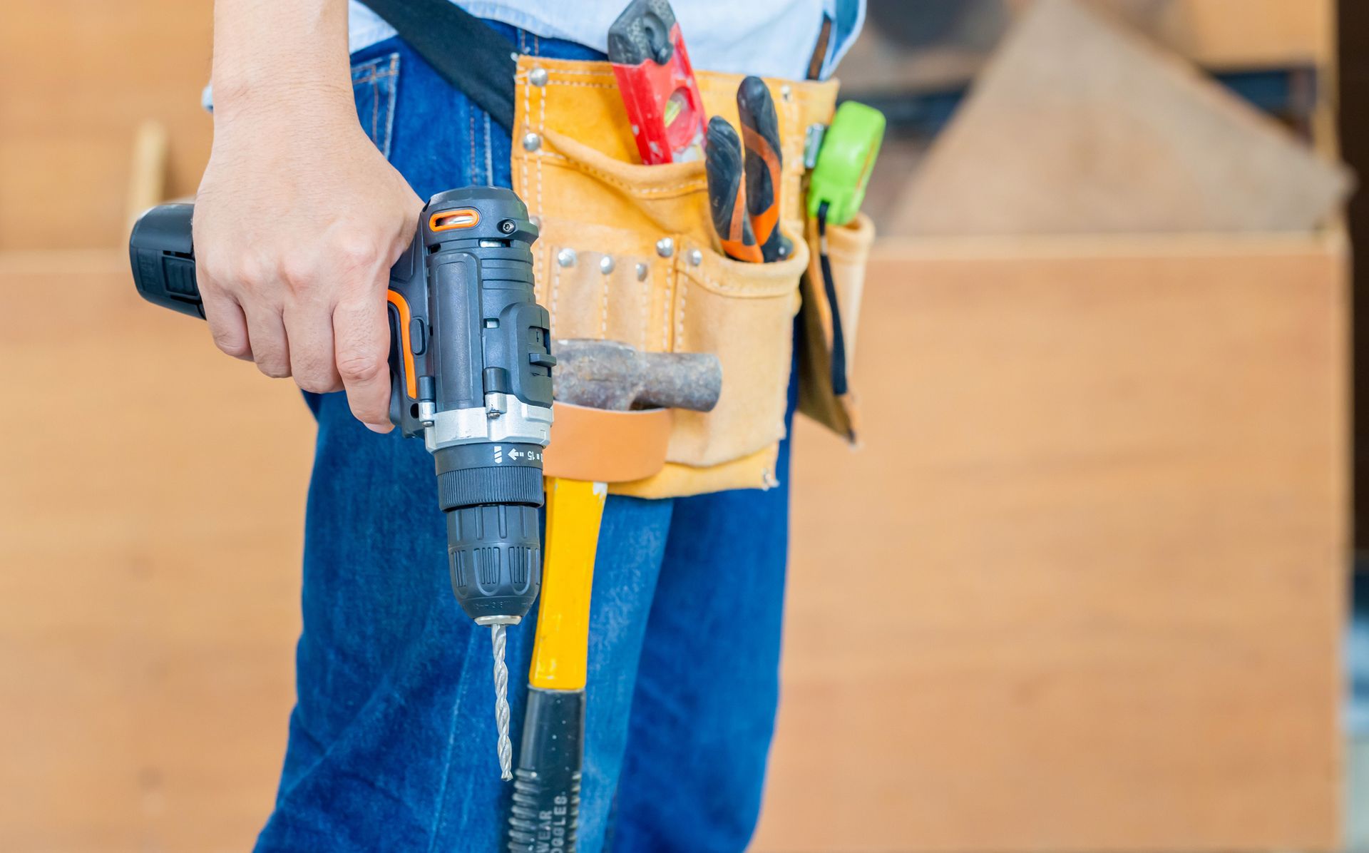 Person in blue jeans holding a drill, wearing a tool belt with a hammer and other tools.