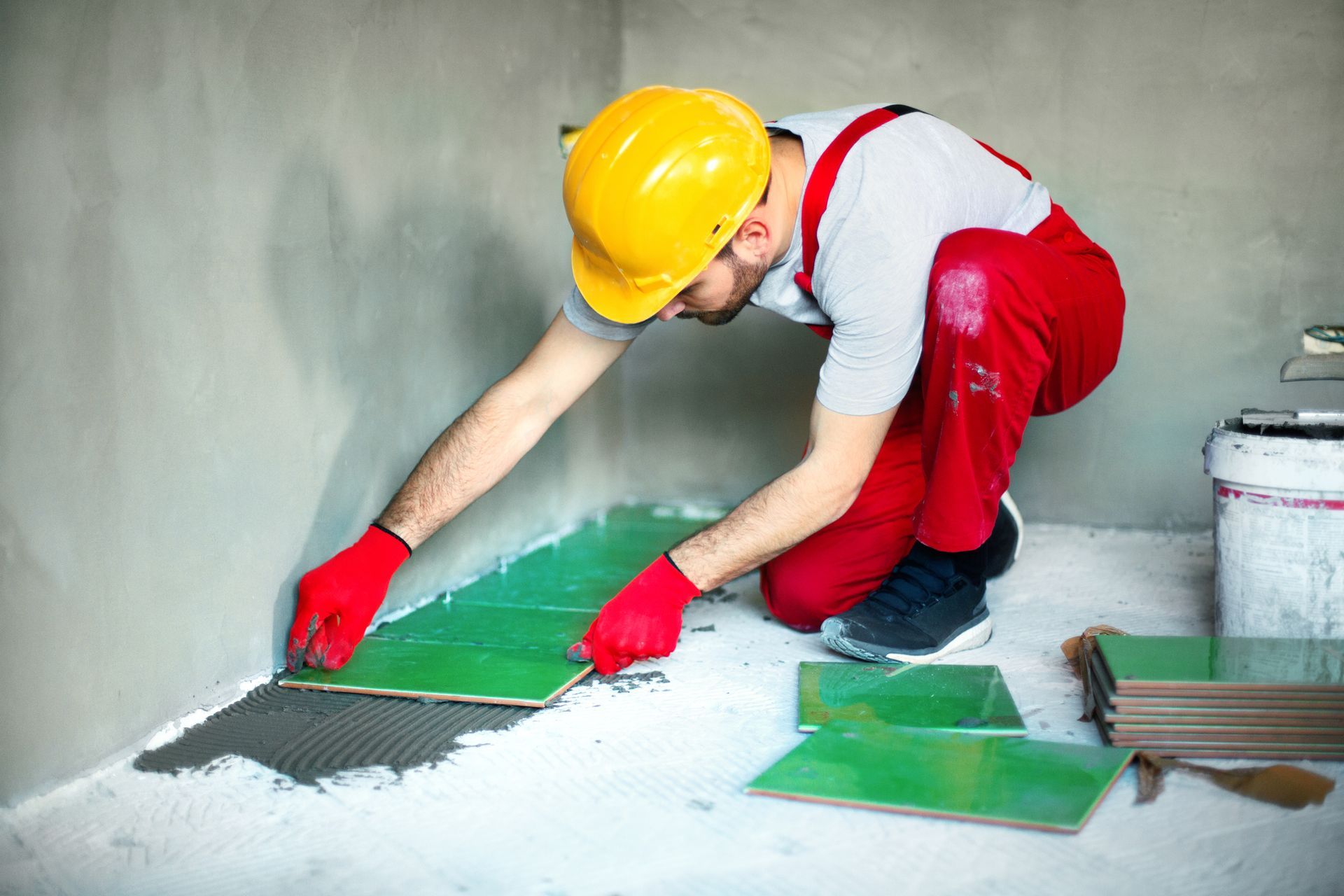 Construction worker laying green tiles in a room; he wears a yellow helmet and red overalls.