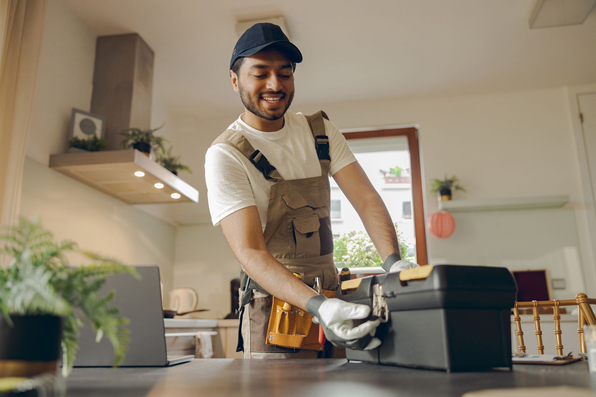 Handyman in overalls opening a toolbox in a bright kitchen. Smiling, wearing gloves and cap.
