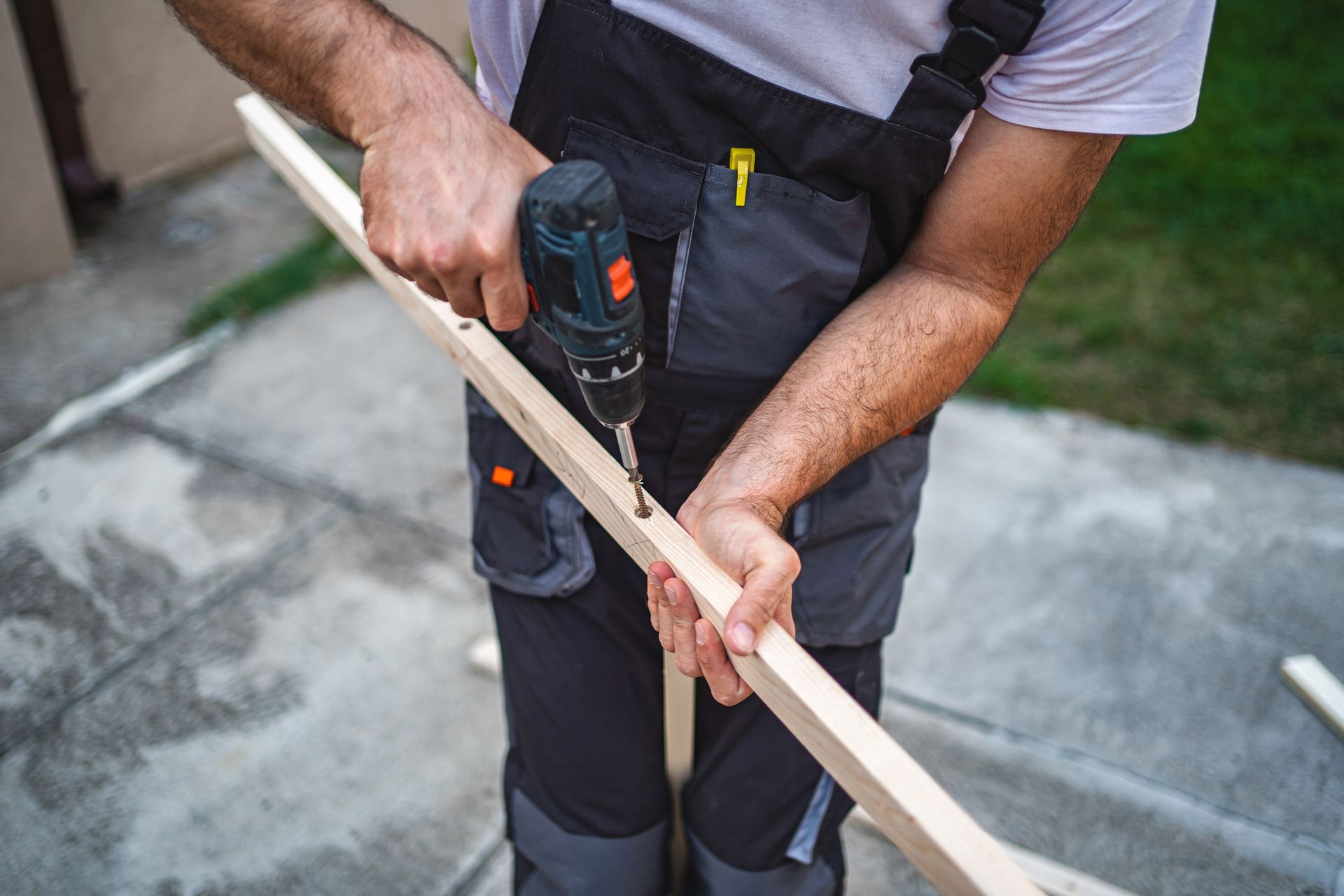 Person in work overalls using a drill on a wooden board outdoors.