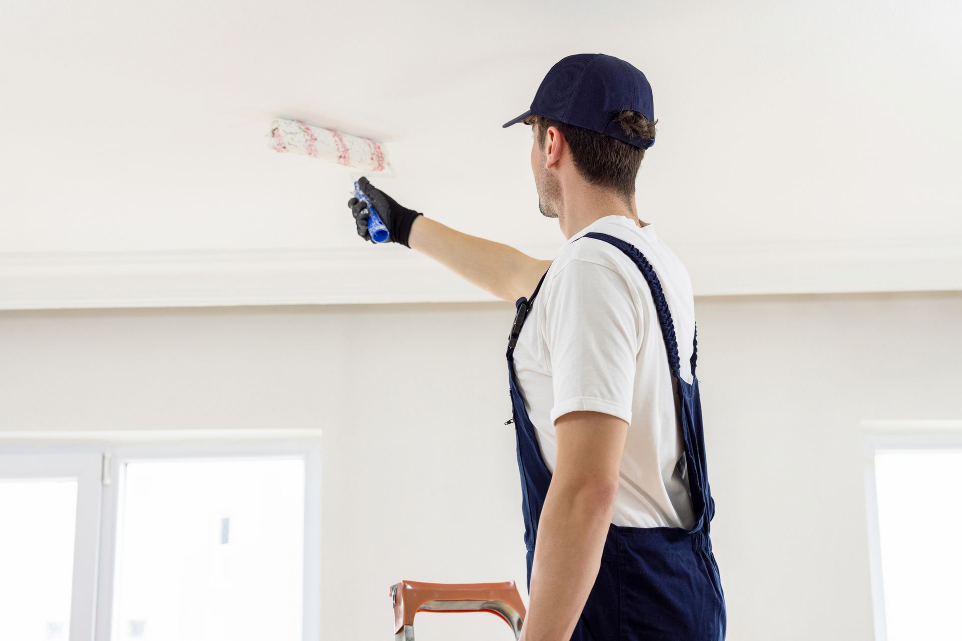 Painter in blue overalls and hat painting a white ceiling, reaching upward.