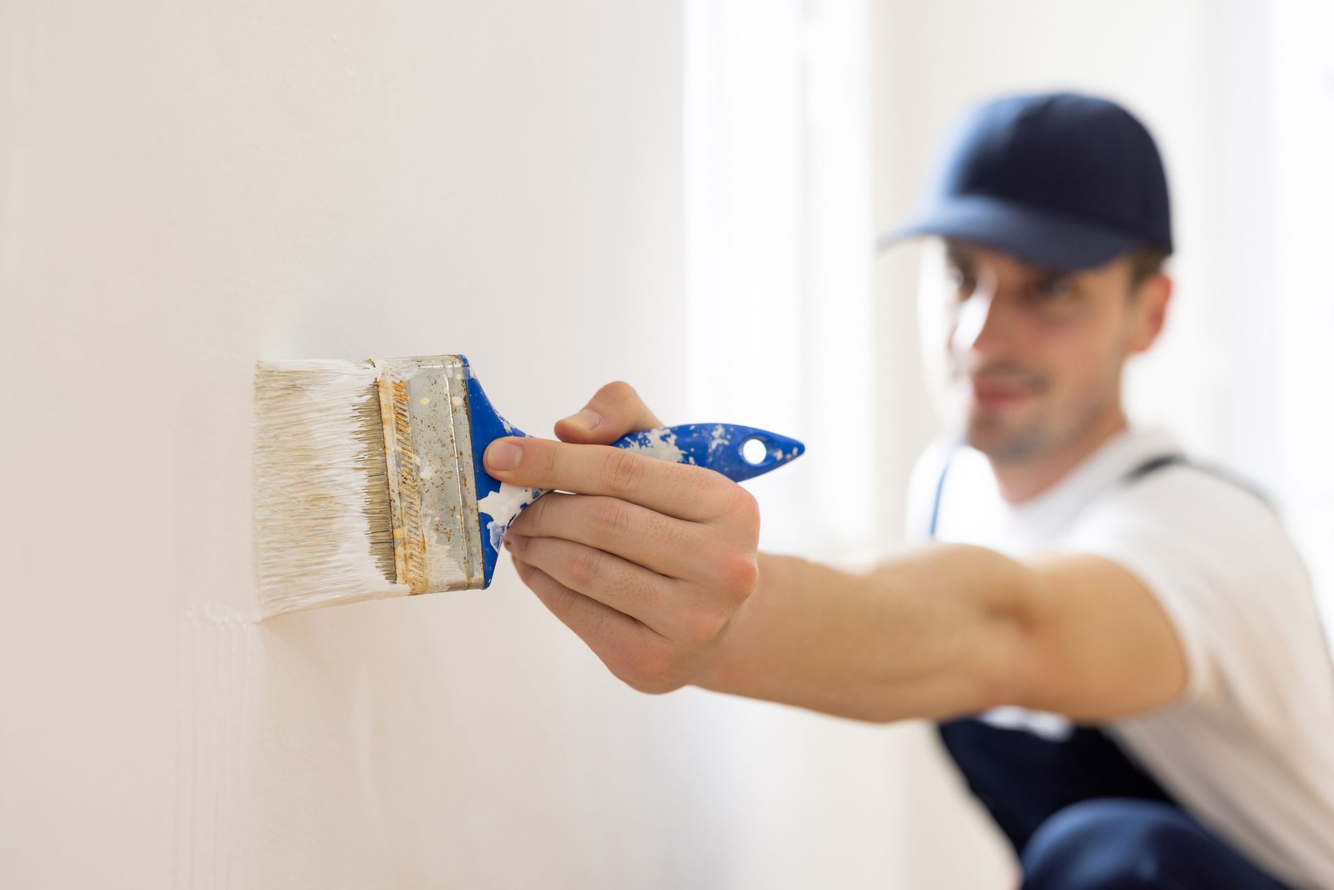 Man in blue cap painting white wall with brush.