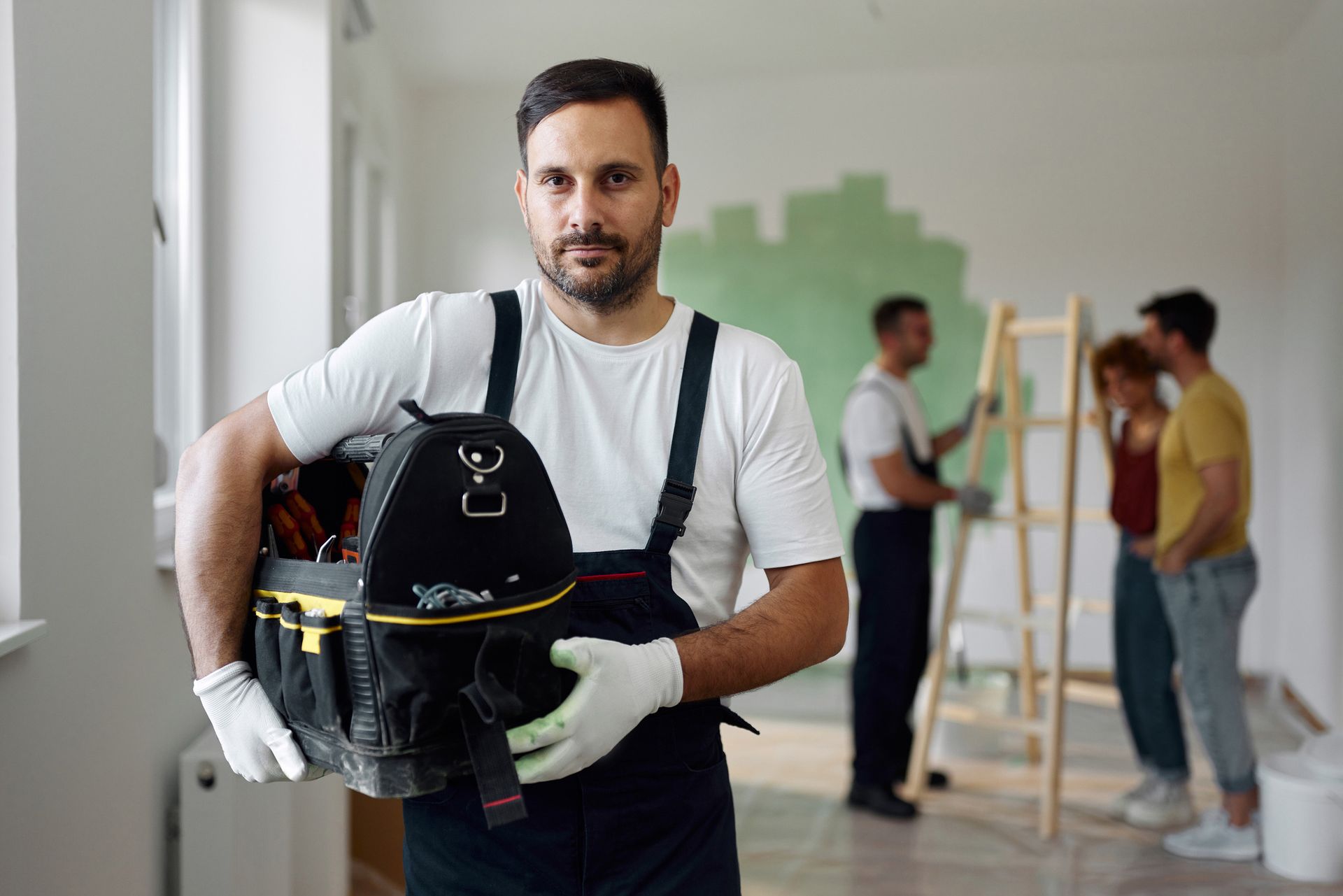 Man in overalls and gloves holds toolbox, looking at the camera. Two workers and couple in background.