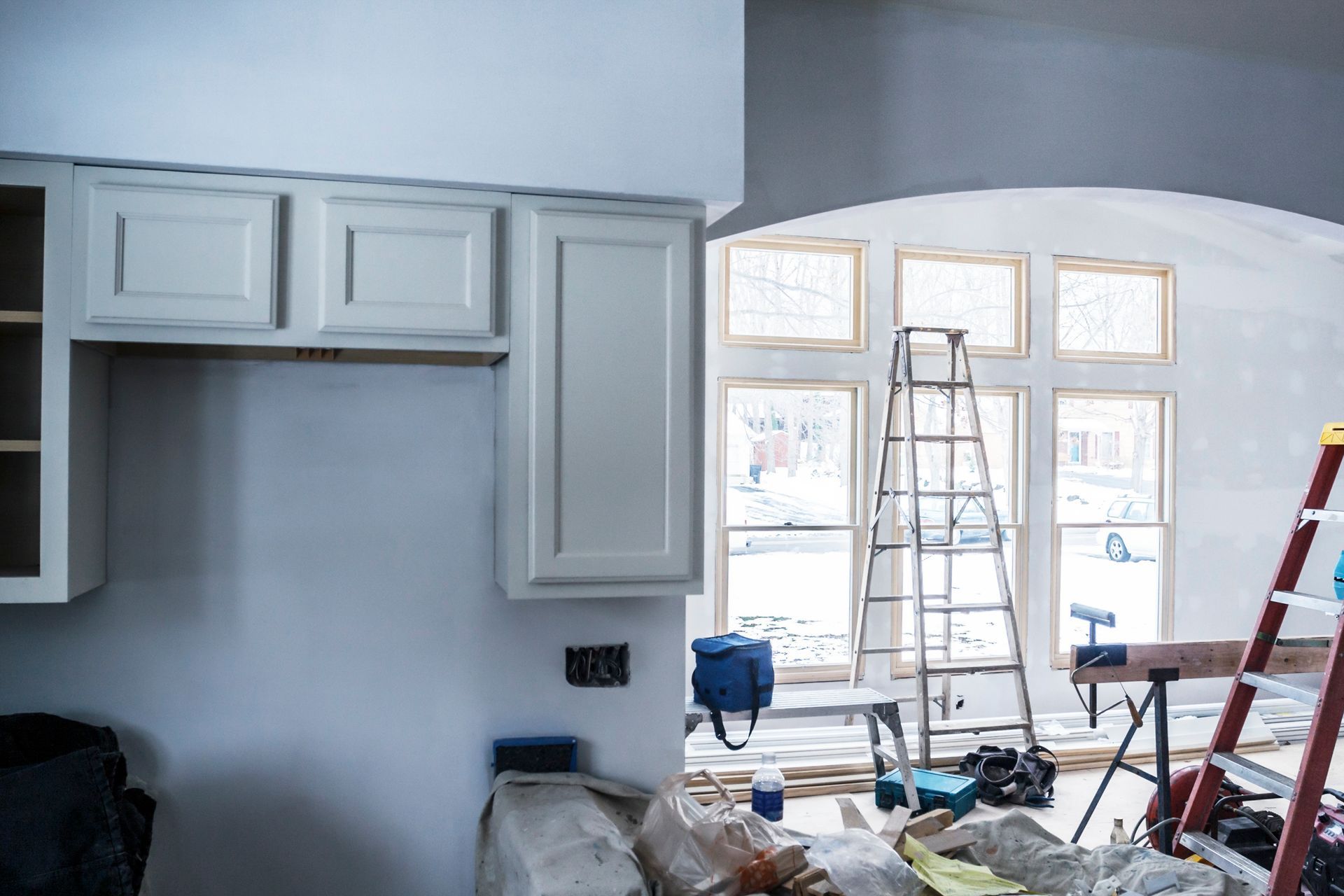Kitchen renovation in progress: white cabinets, arched windows with ladder, construction materials.