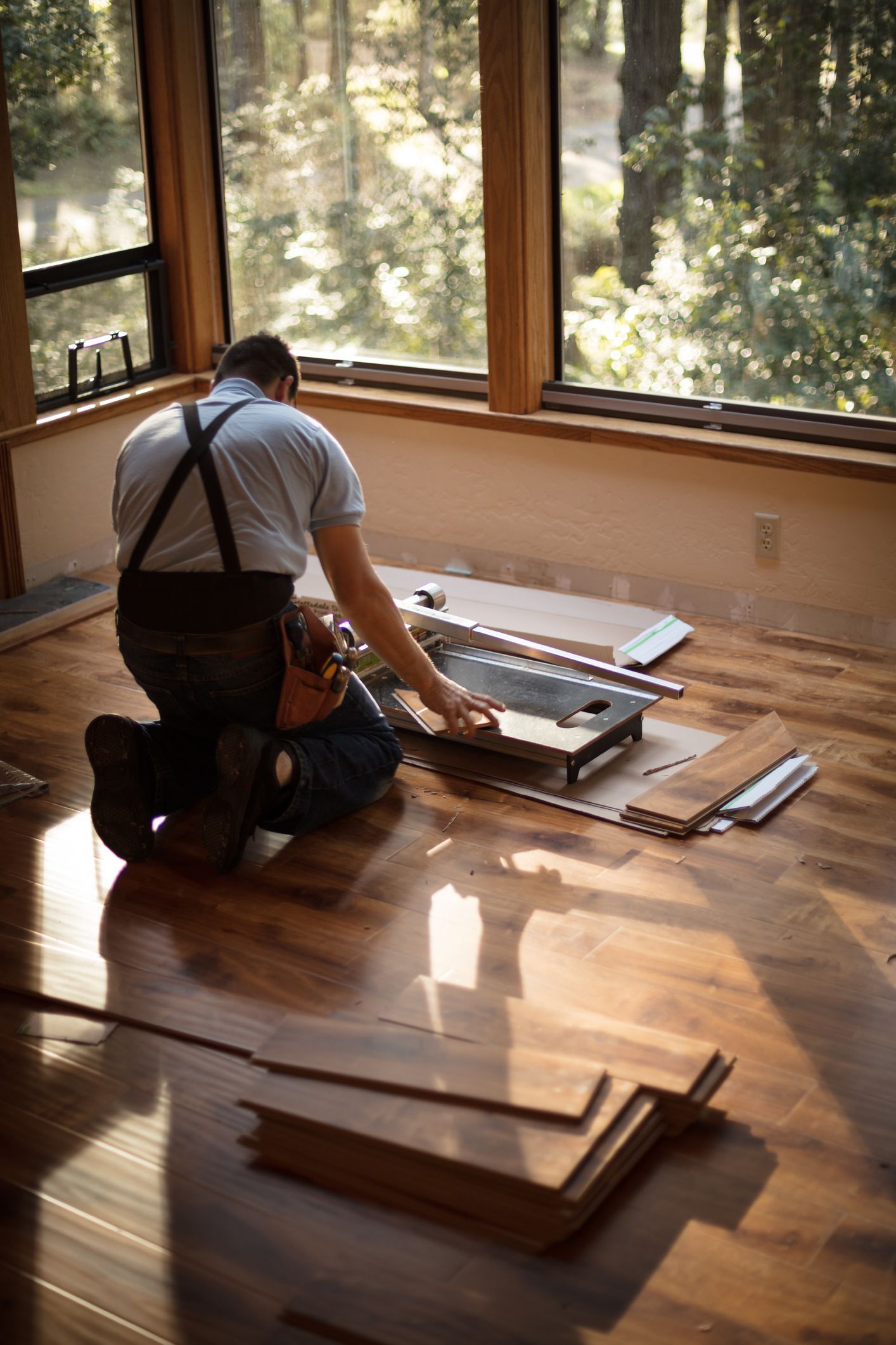 Man kneeling, installing wood flooring in a room with large windows, trees visible outside.