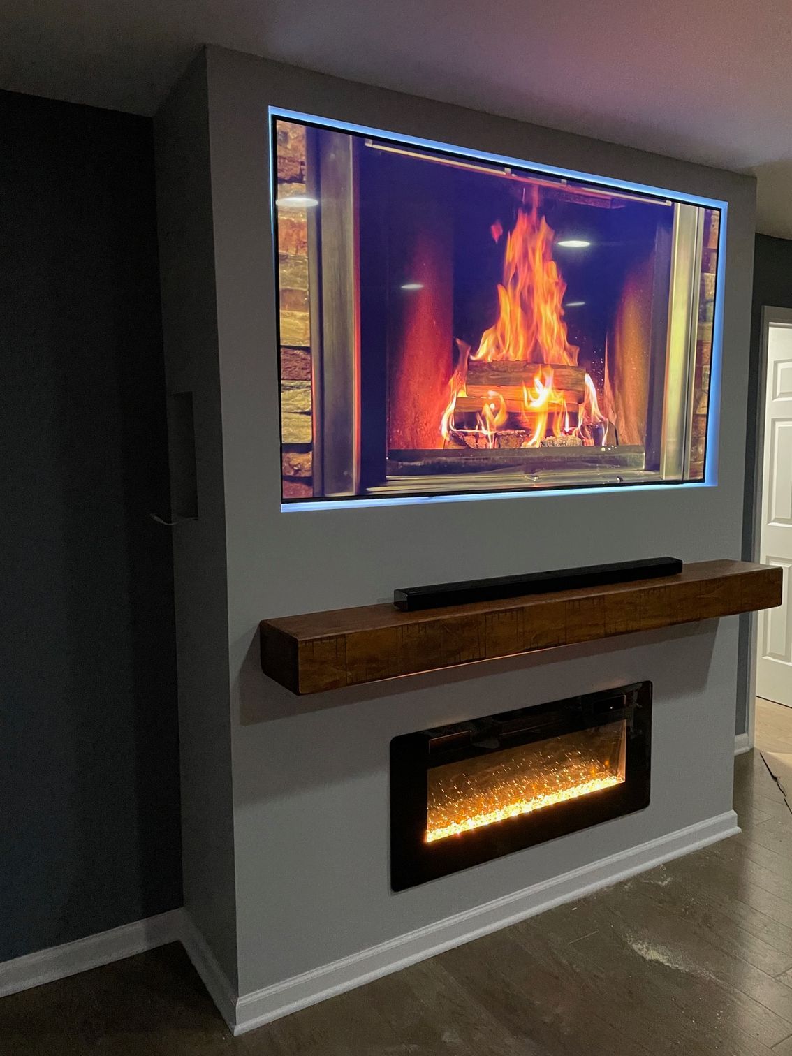 Gray wall with TV above electric fireplace, wooden mantel, and flames on screen.