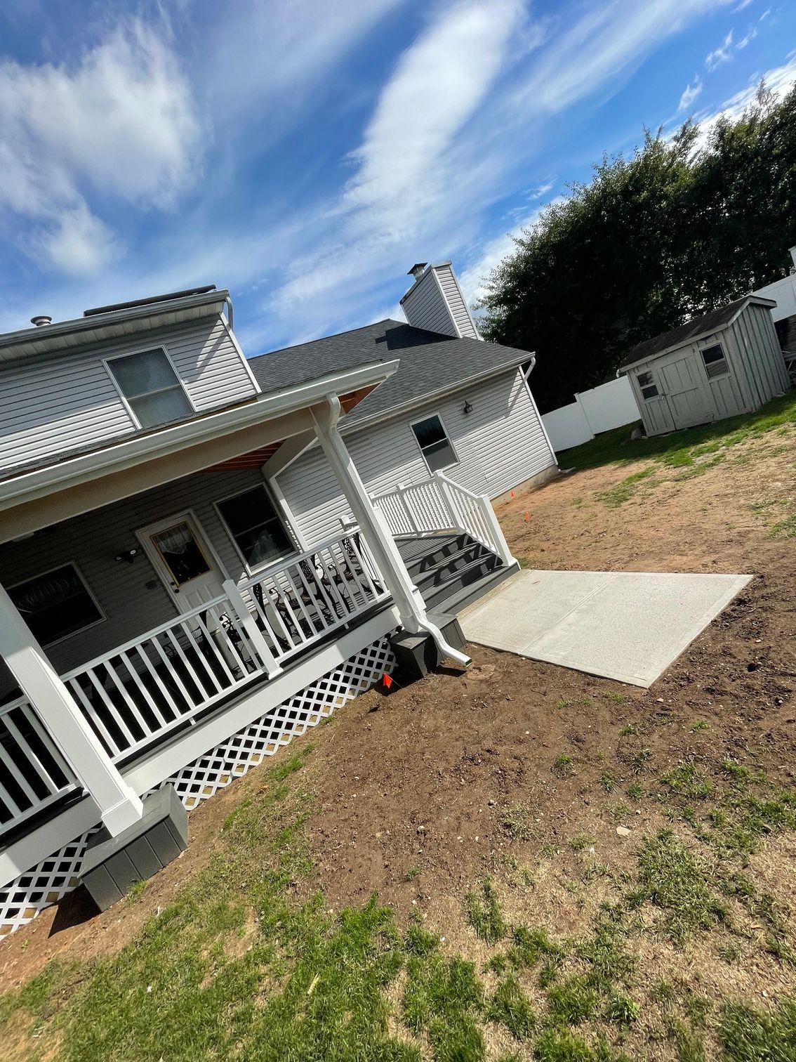 Backyard with a two-story gray house, patio, shed, and blue sky. Brown and green grass.