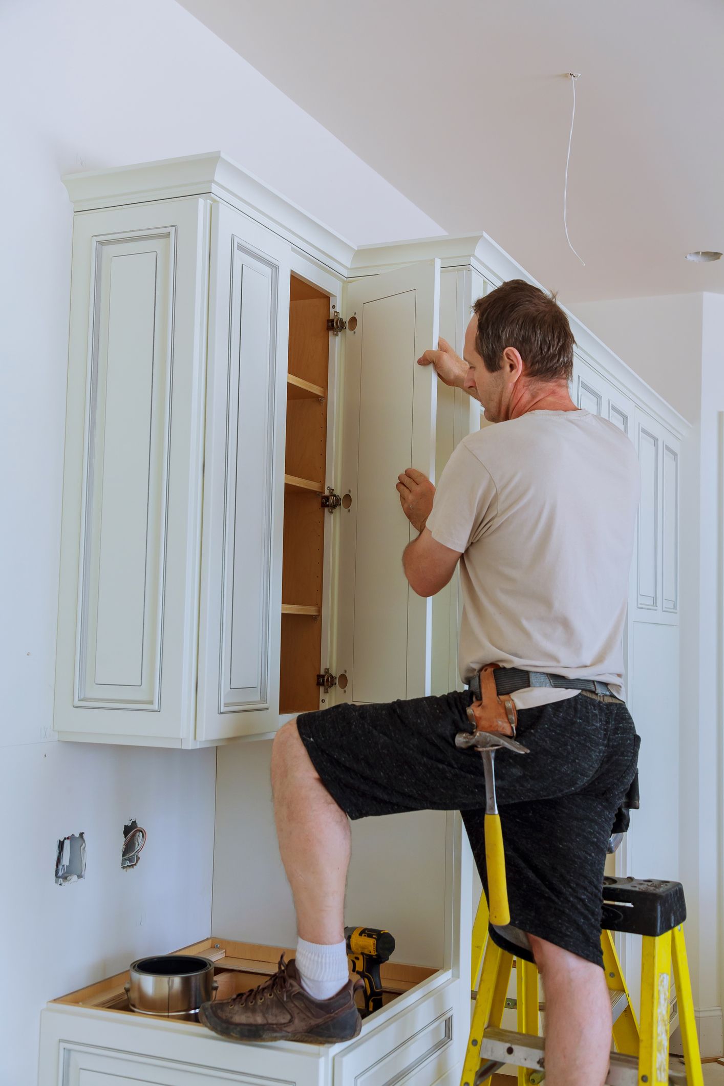 A man is kneeling on a ladder working on a kitchen cabinet.