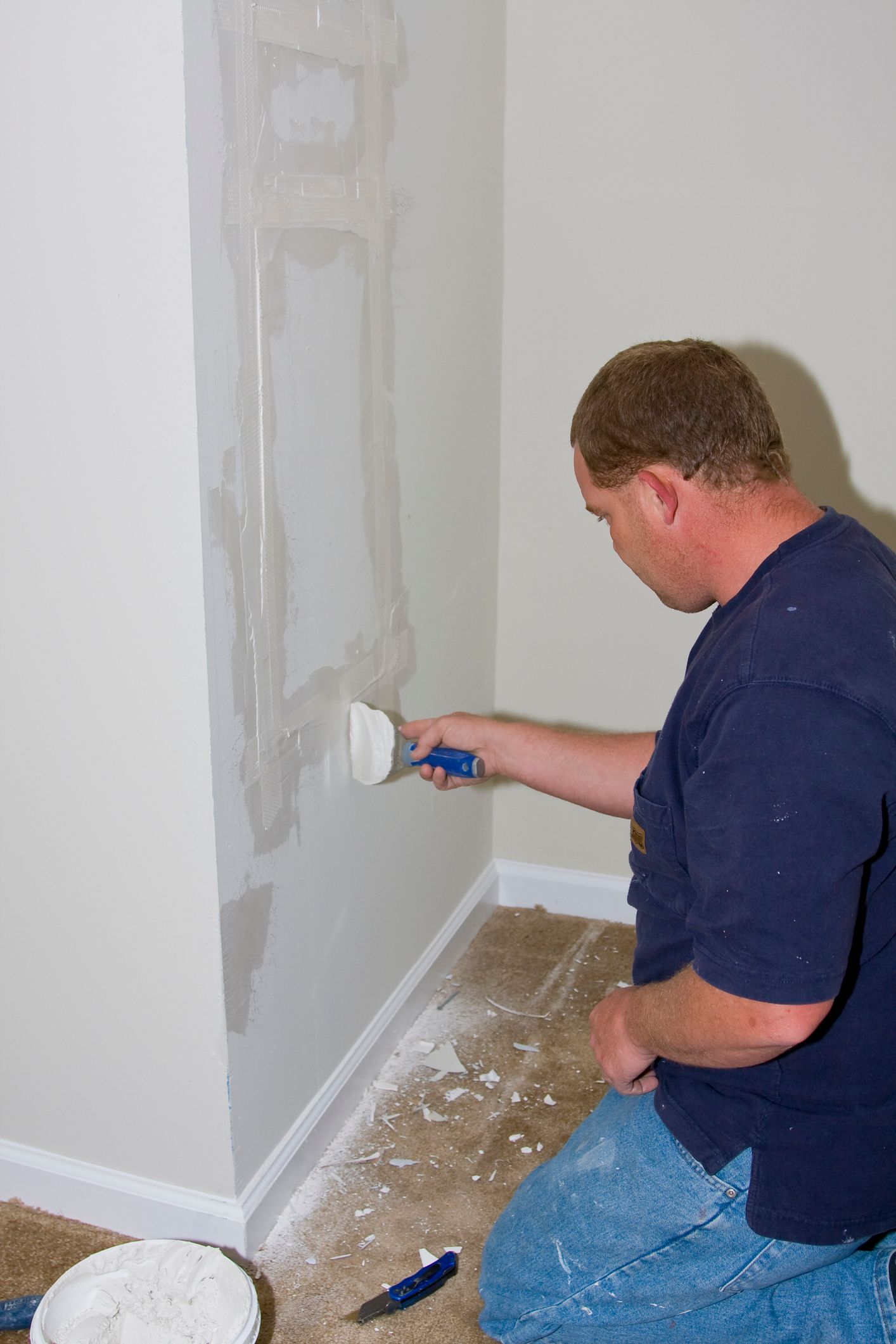 Man kneeling, applying spackle to a wall with a putty knife. White spackle covers the wall in a room.