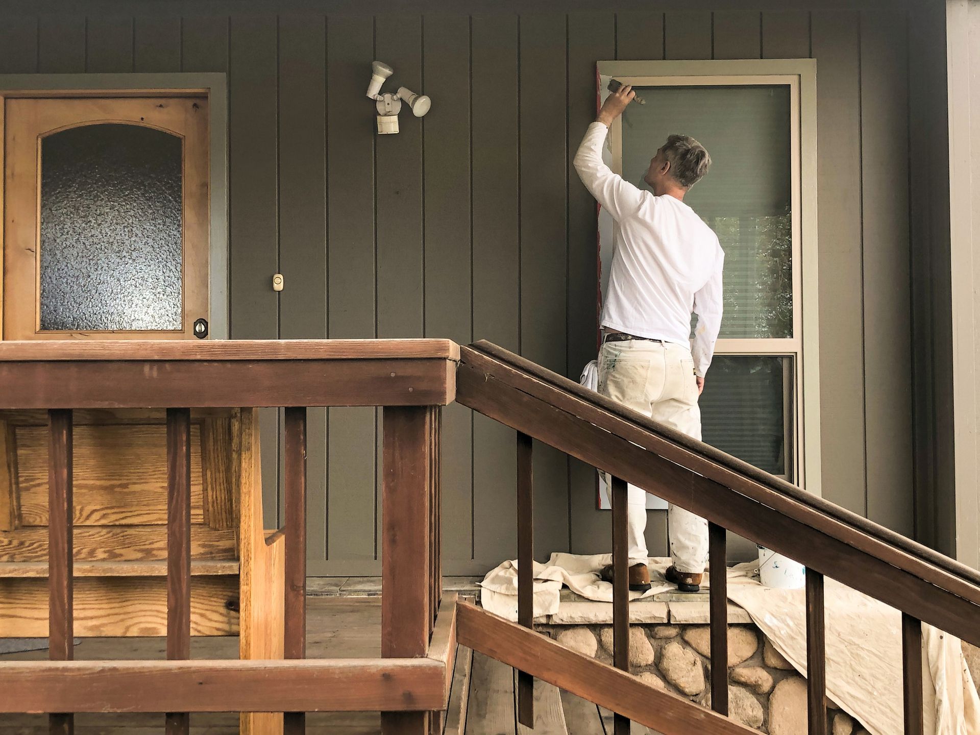 Man painting window trim on a house with brown siding and a porch.