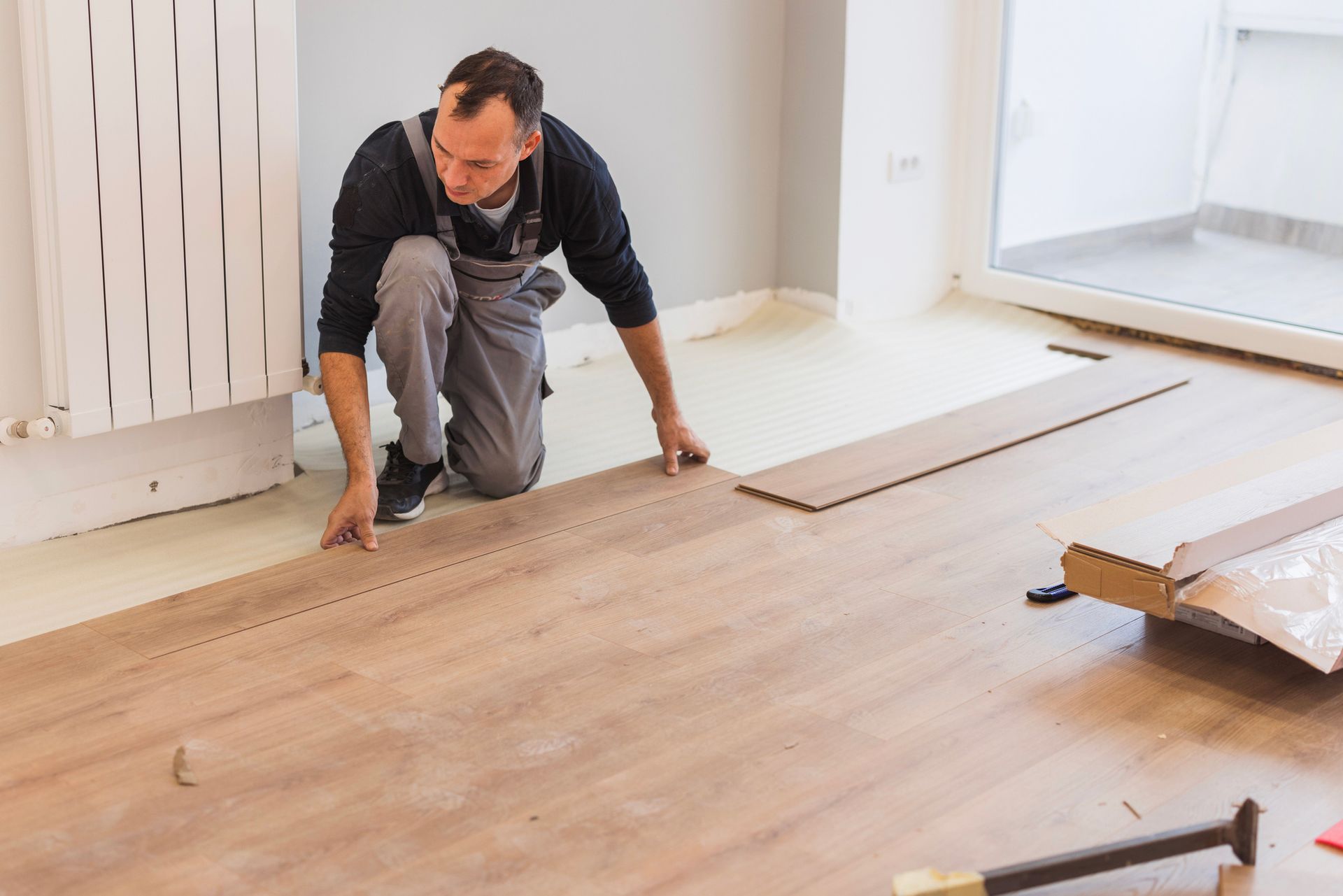 Man kneeling, installing wooden floorboards in a room.  He wears work clothes.