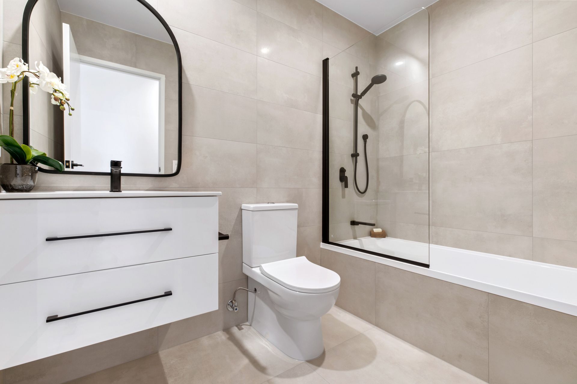 Modern bathroom with white toilet, vanity, and bathtub, beige tile walls, black accents.