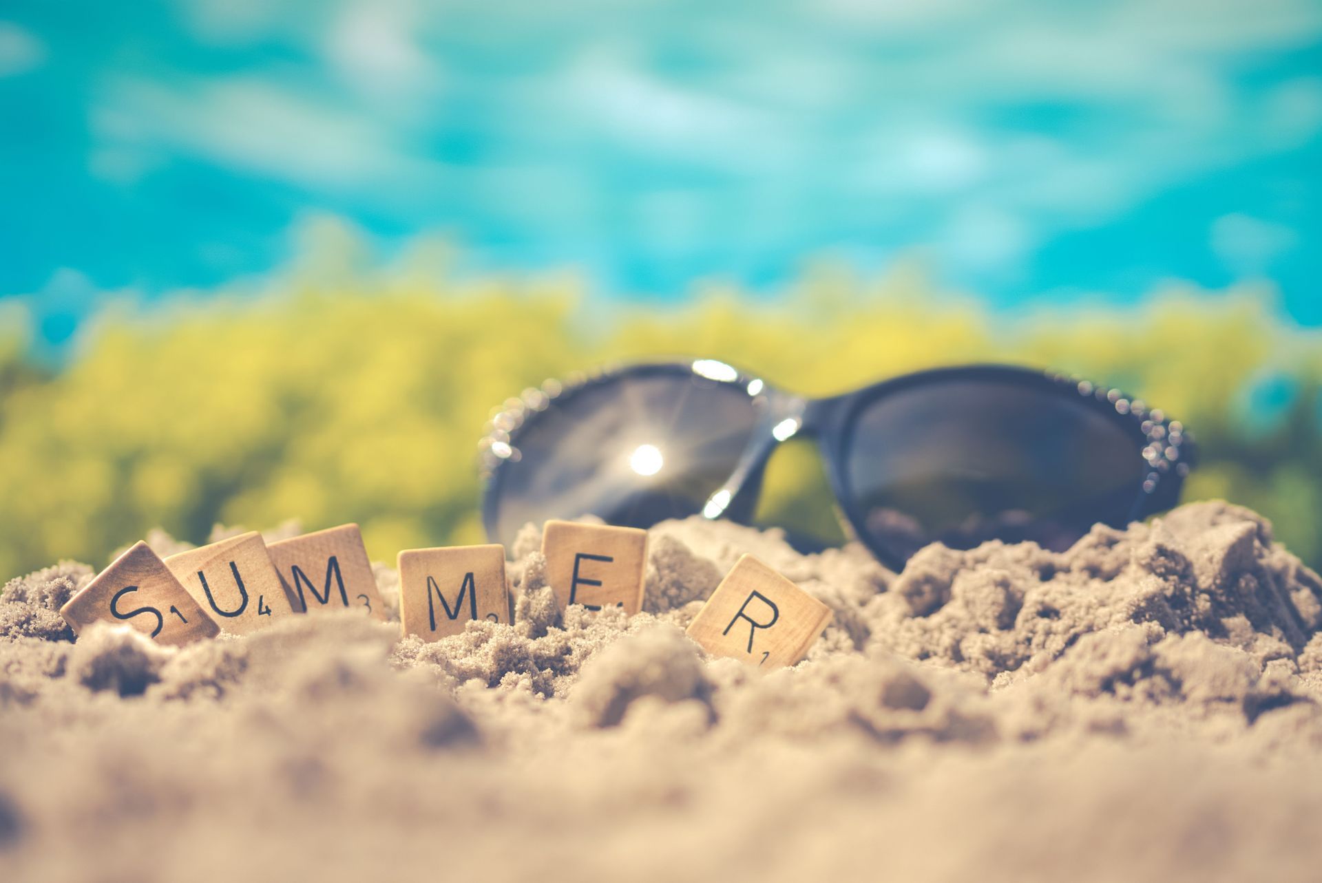 Sunglasses on sand with wooden letters spelling “SUMMER” against a blue sea background