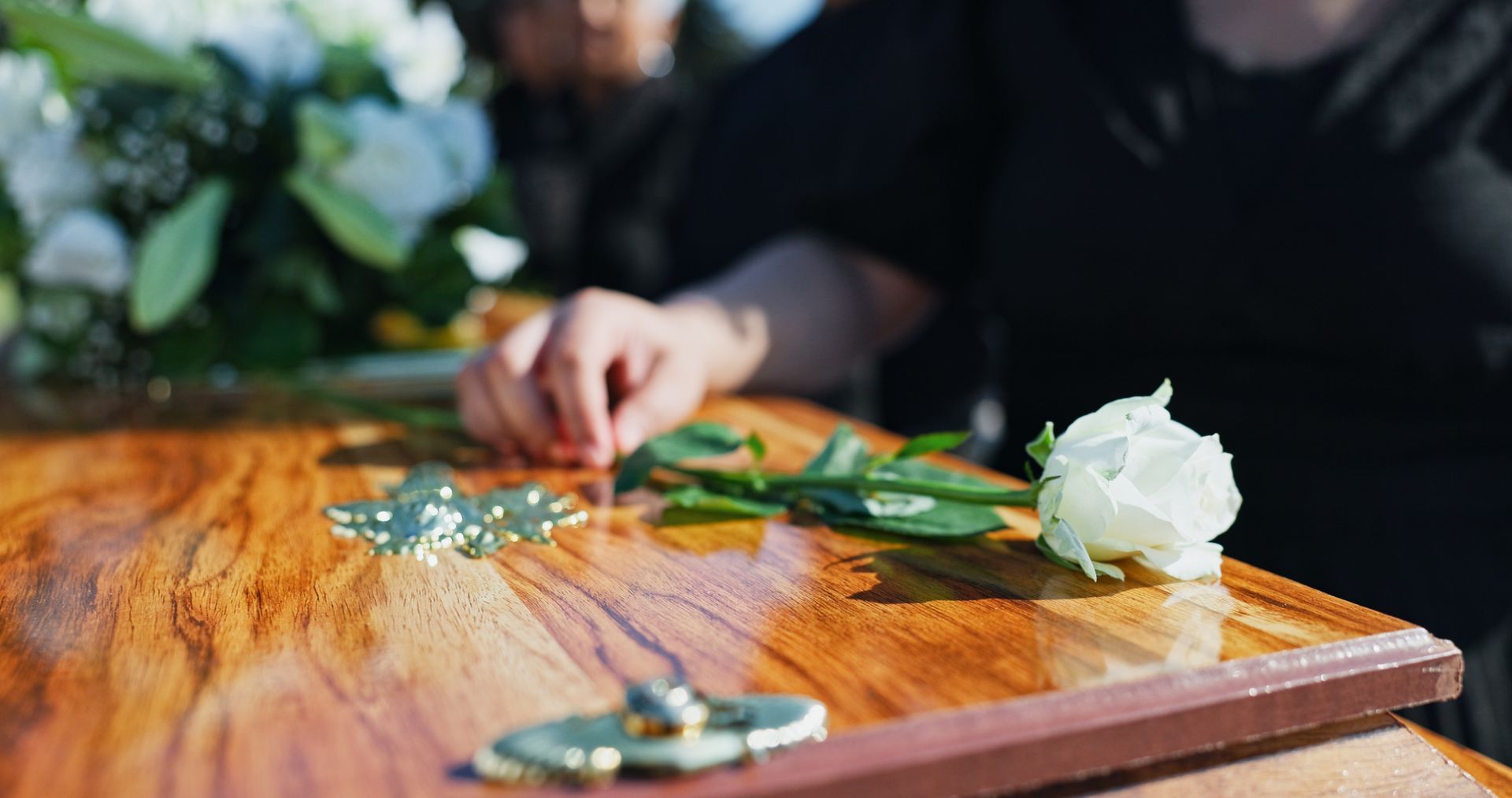 Close-up of hand touching a white rose on a polished wooden casket. Mourner in black clothing.