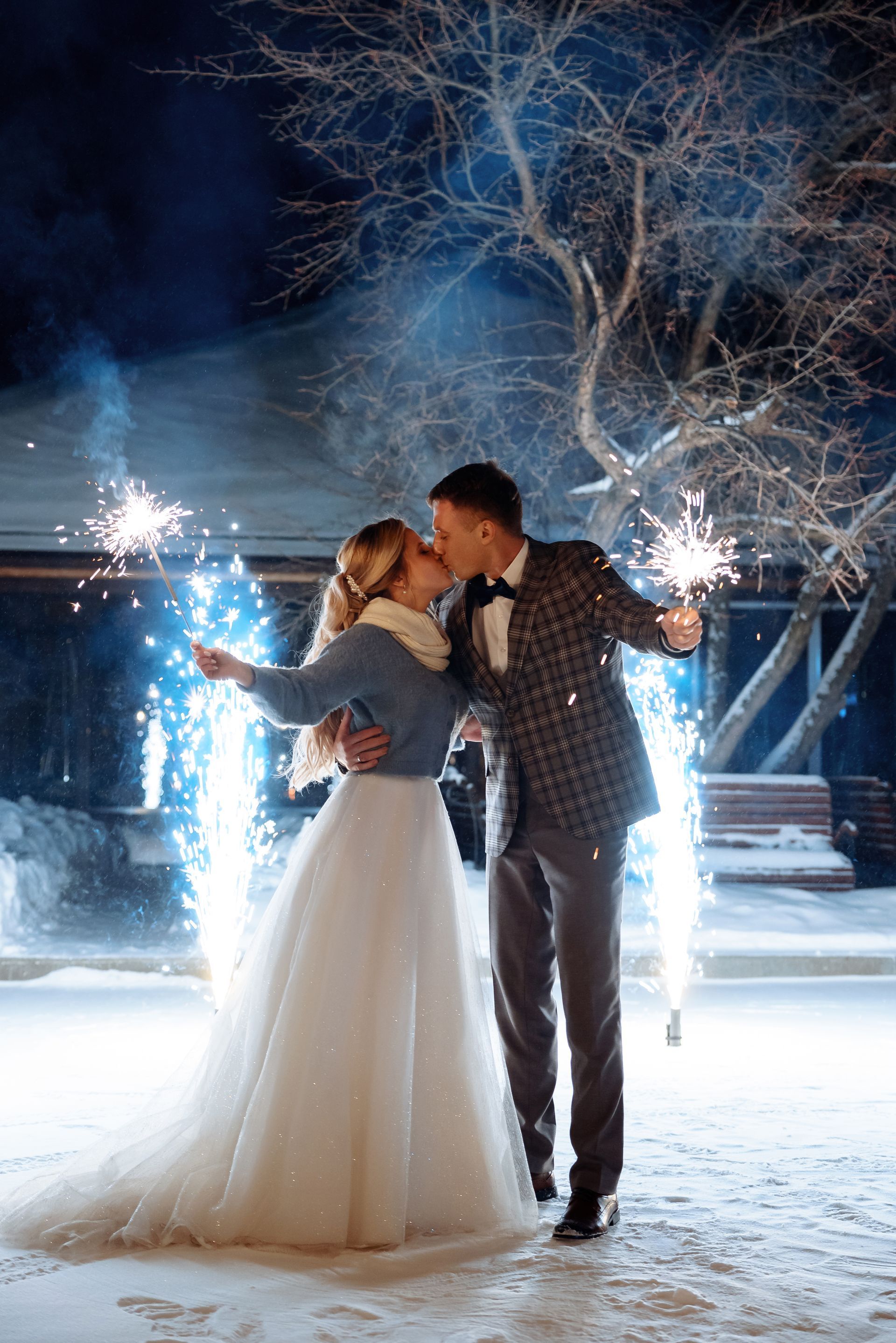 A bride and groom are kissing in the snow while holding sparklers.