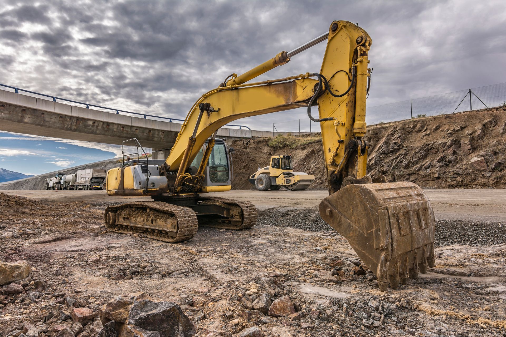 Escavatore giallo in un cantiere con un ponte sullo sfondo.