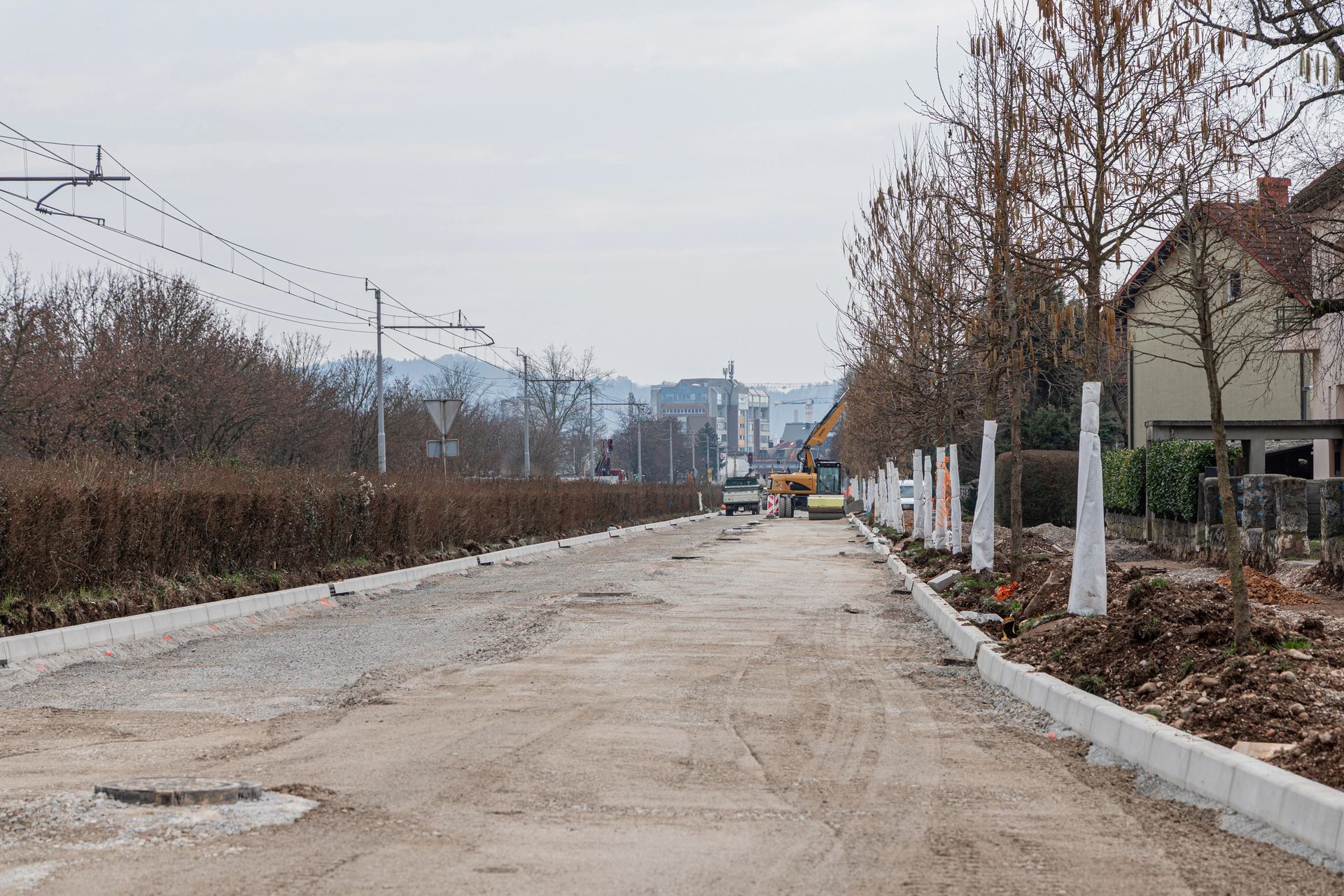 Scena di lavori stradali con terra, ghiaia e mezzi da cantiere. Alberi ed edifici costeggiano la strada.
