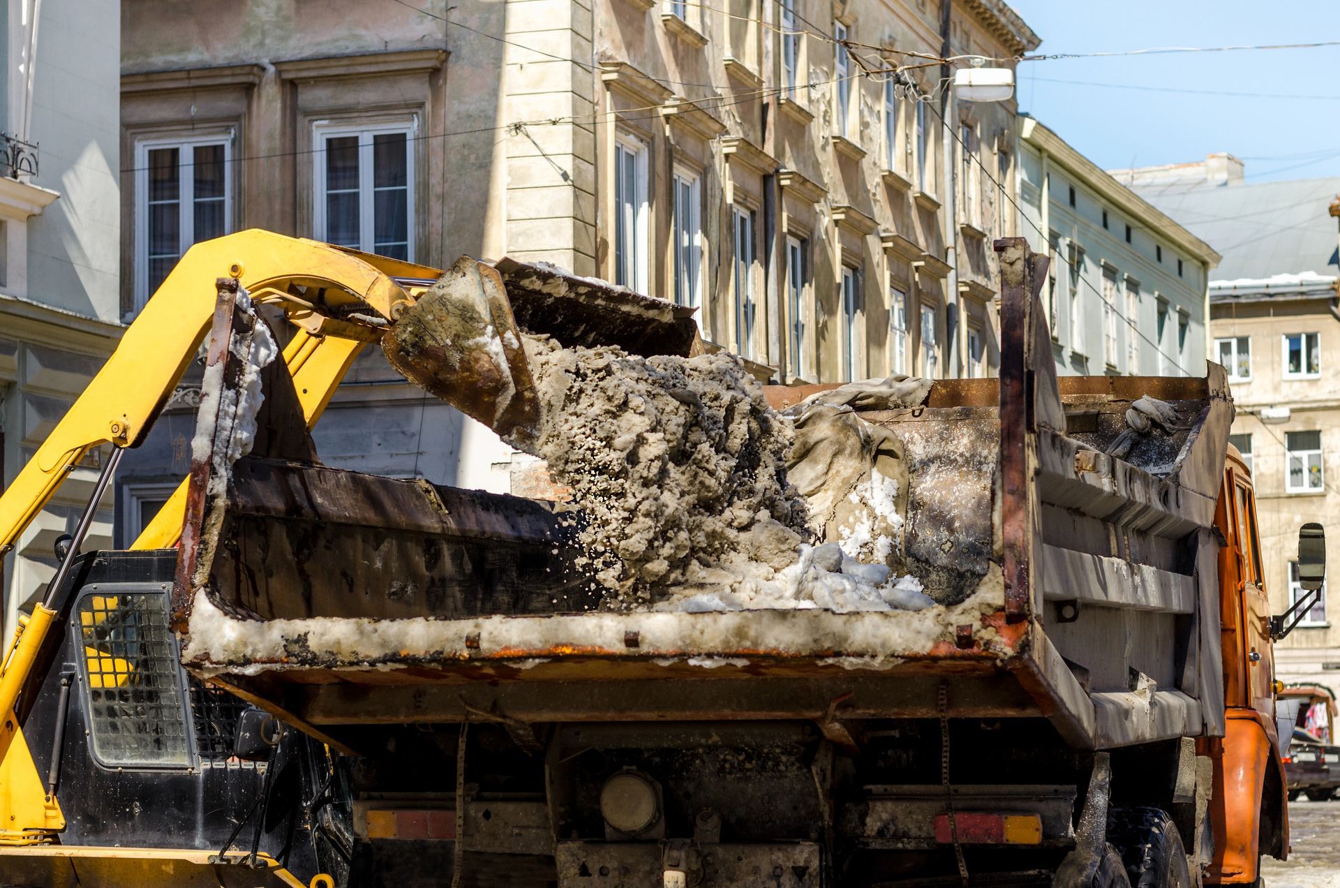 Un escavatore giallo carica detriti su un autocarro con cassone ribaltabile in una strada cittadina.
