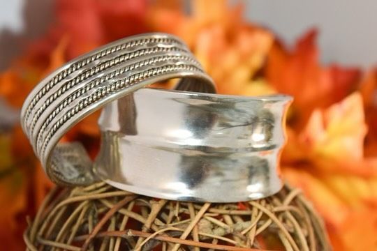 Two silver bracelets stacked, one rope design, one flared, on a woven ball with orange leaves.