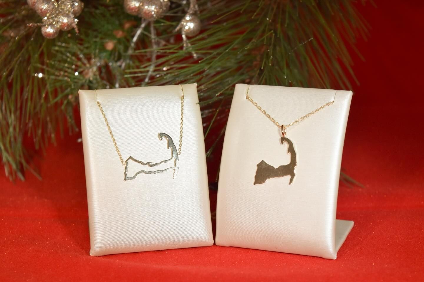 Two gold necklaces shaped like Cape Cod displayed on white stands, with a Christmas background.