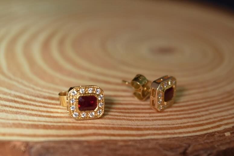 Gold stud earrings with a red center stone, surrounded by small diamonds, resting on a wood surface.