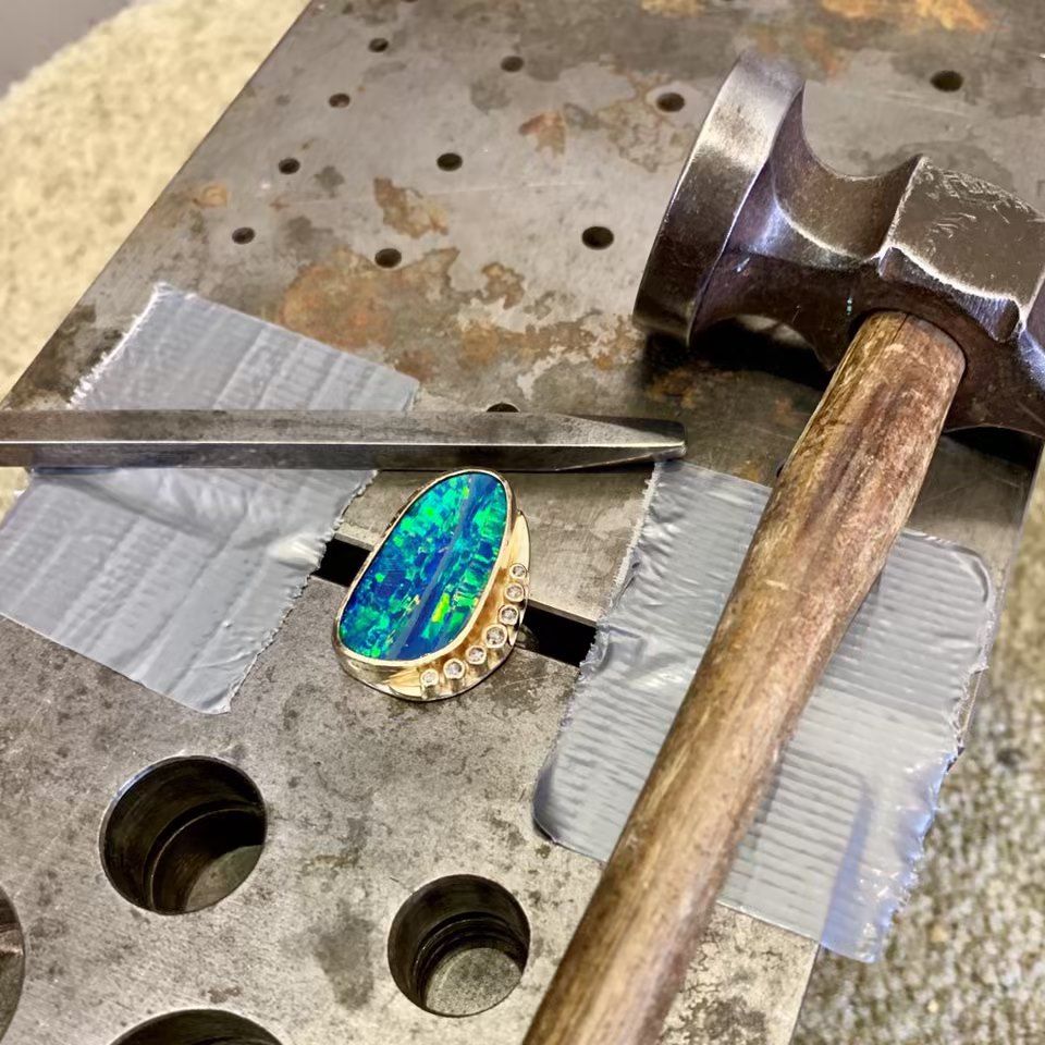 Ring with opal gemstone on a metal workbench with hammer and chisel.