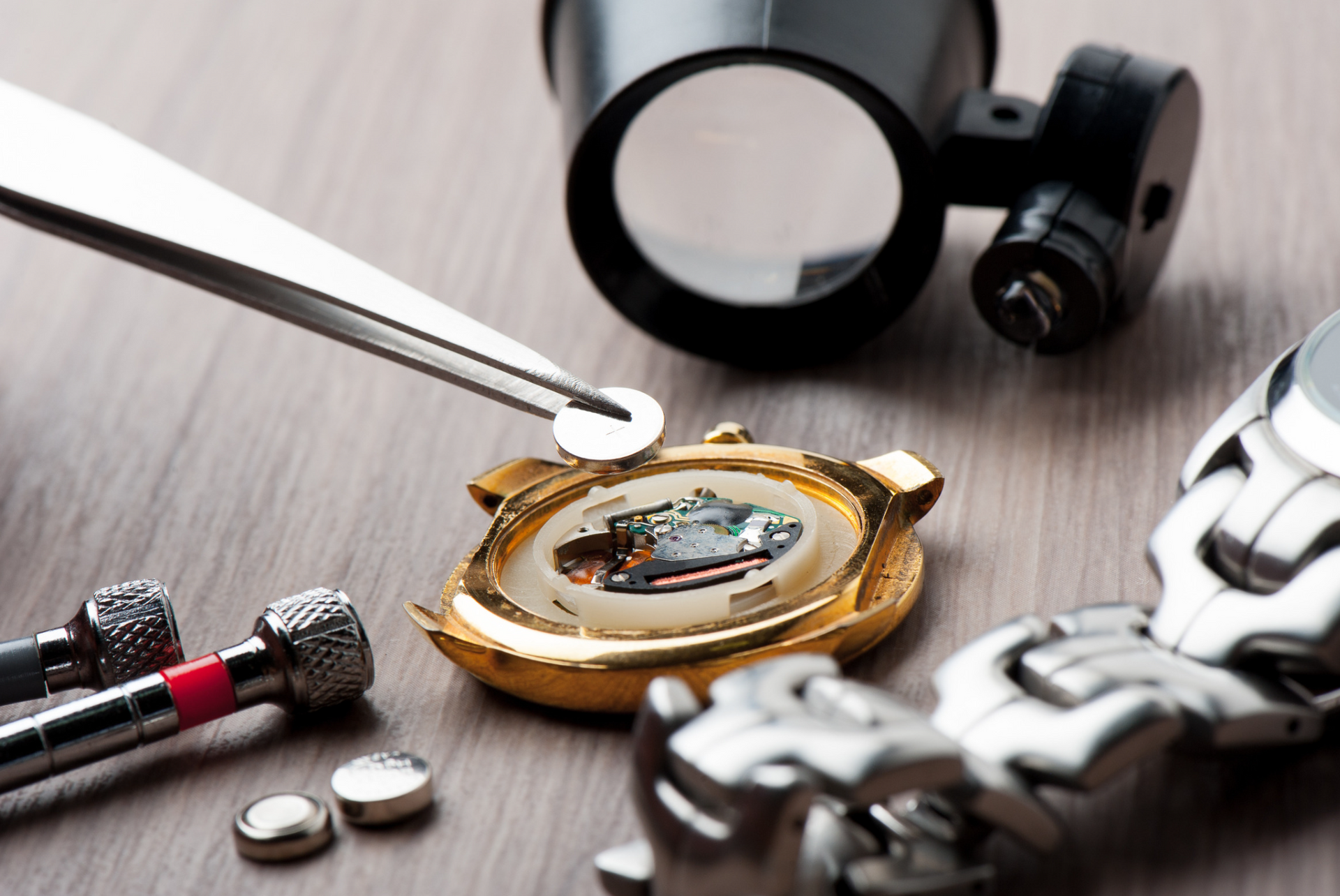 Watch repair: tweezers holding a battery near an open watch, magnifying glass, tools on wooden surface.